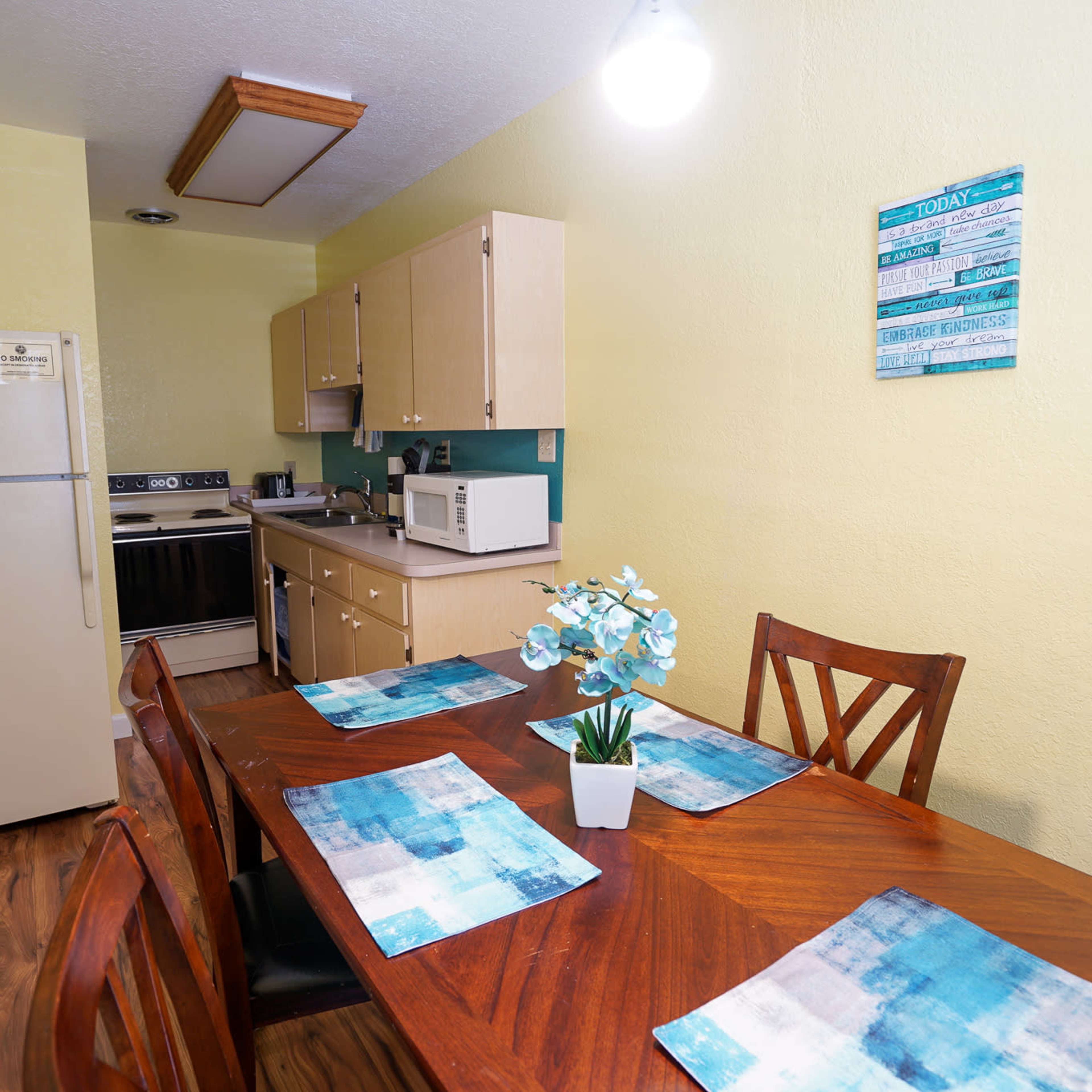A small, functional kitchen with a dining table set for four, featuring a fridge, stove, and microwave against a light green wall.