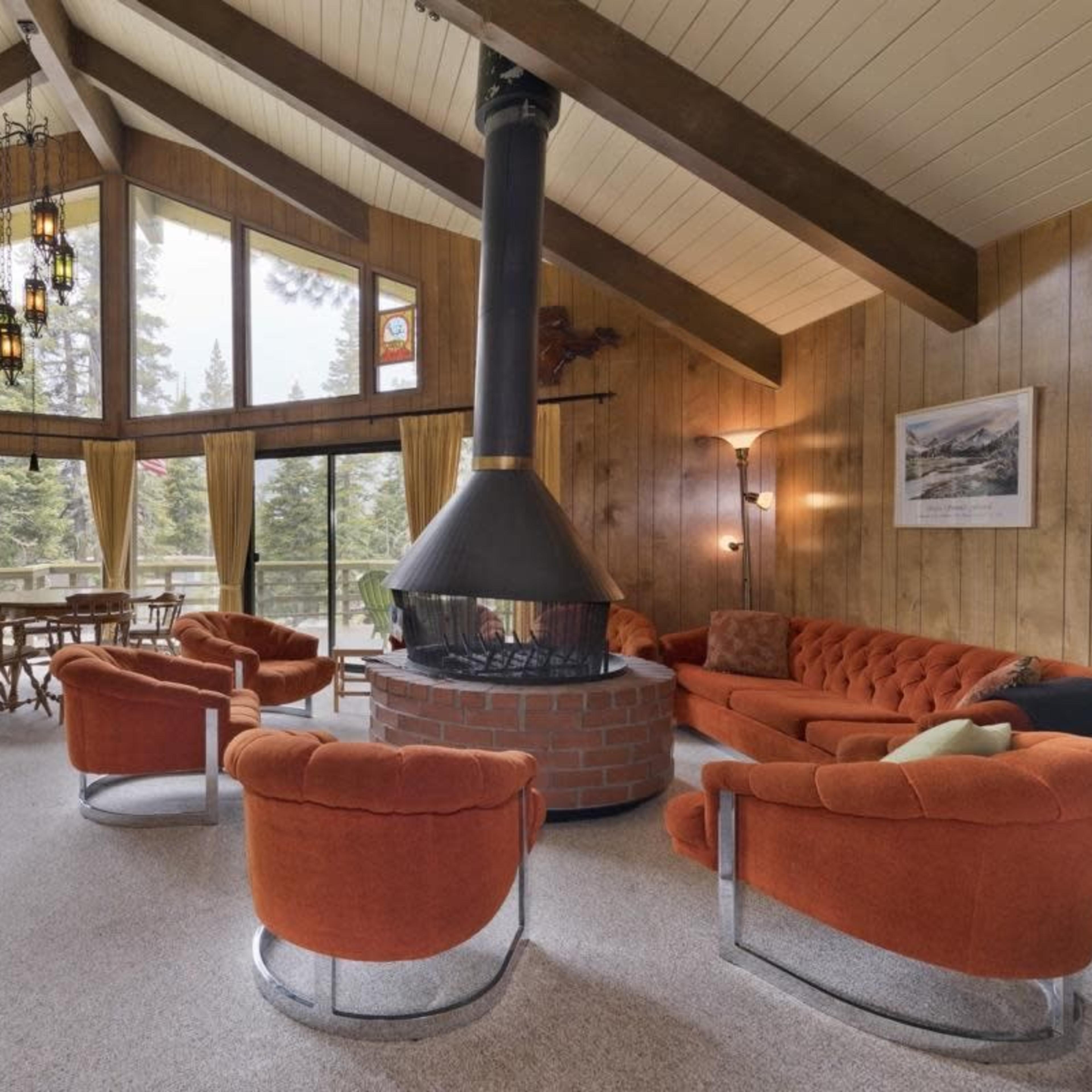 A cozy living room features a central circular fireplace, surrounded by orange furniture and large windows that provide views of the trees outside.