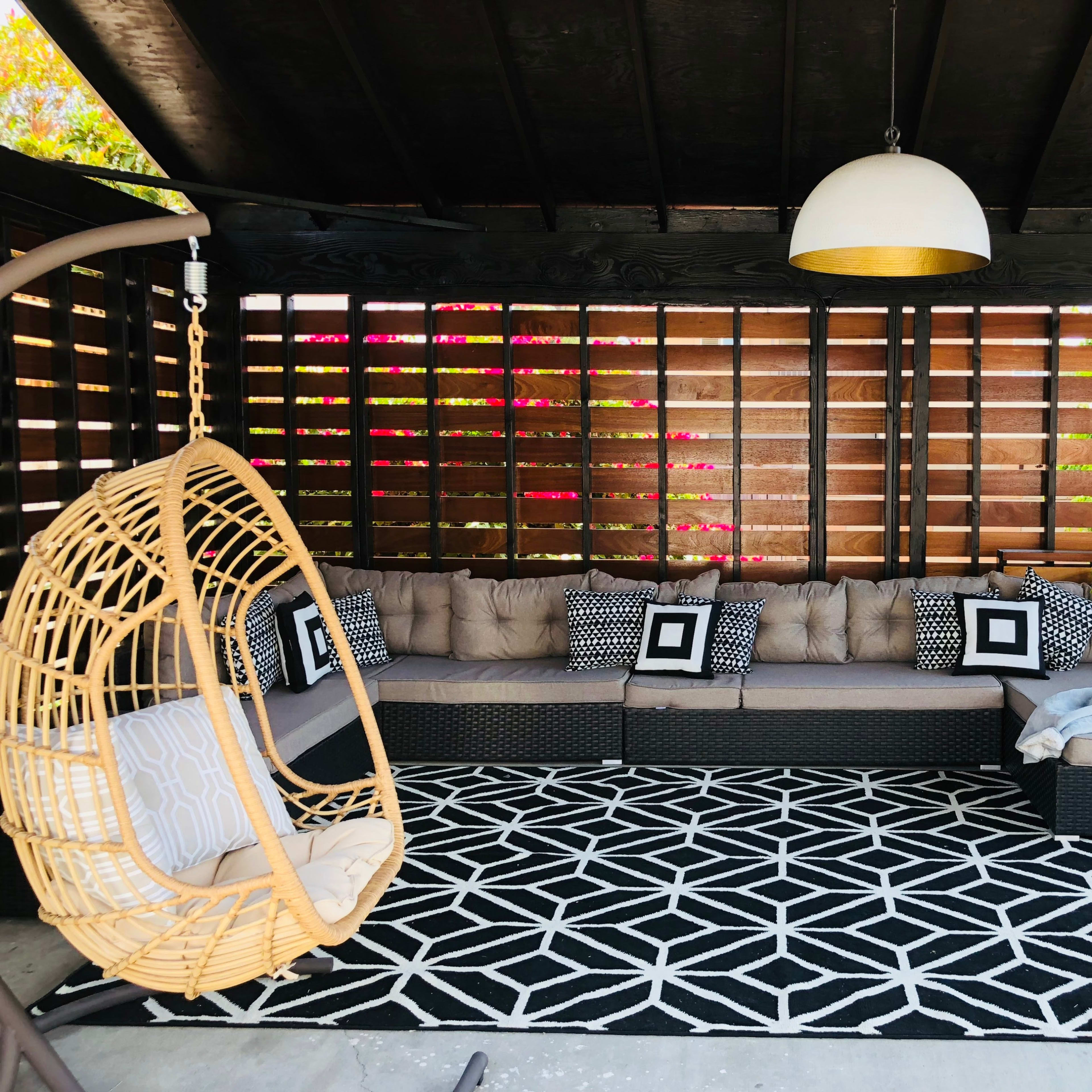 A shaded outdoor seating area with a sectional couch, decorative cushions, a hanging chair, and a geometric-patterned rug.