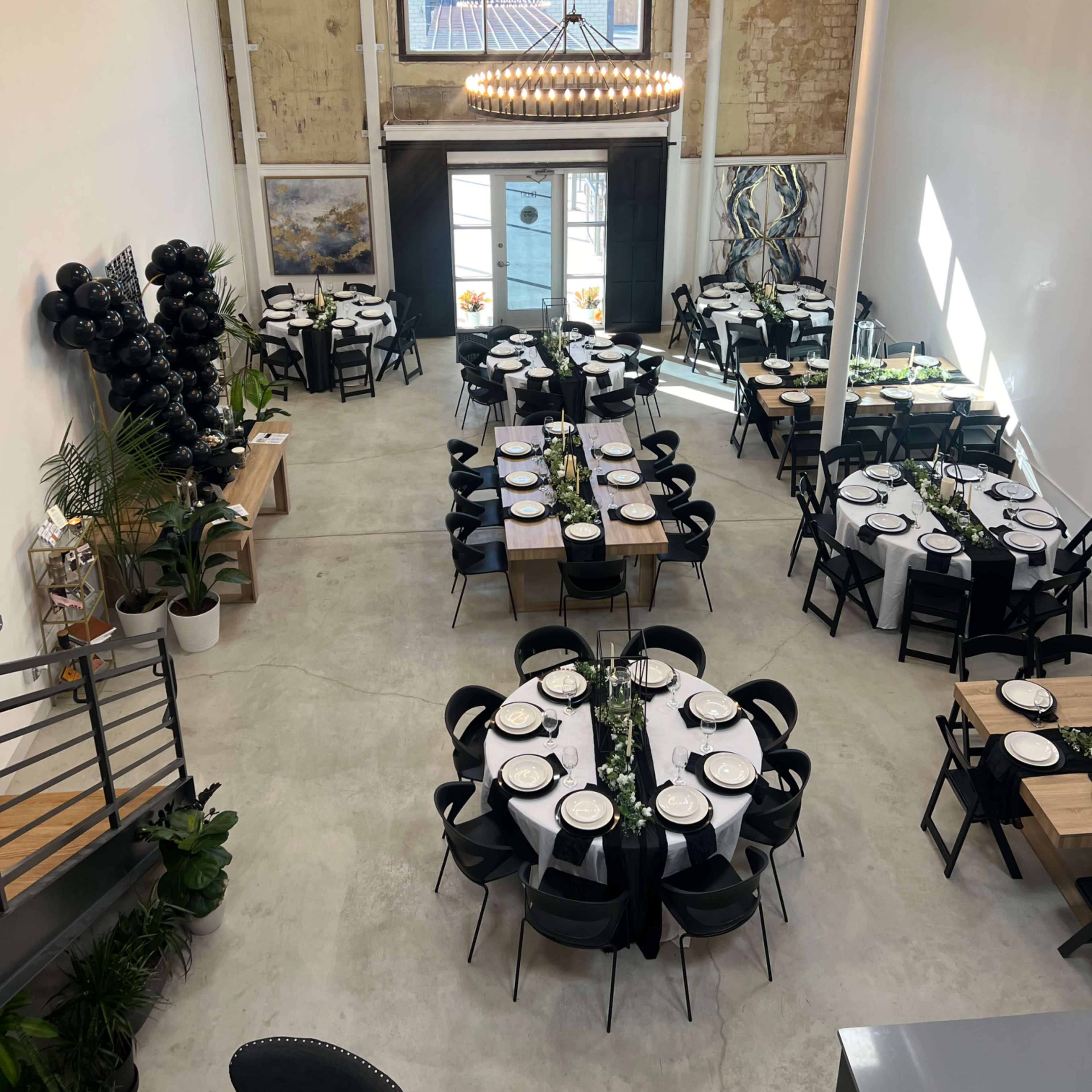 A spacious dining area with two round tables and several rectangular tables set for a formal event, featuring black chairs and a mix of decorative plants.