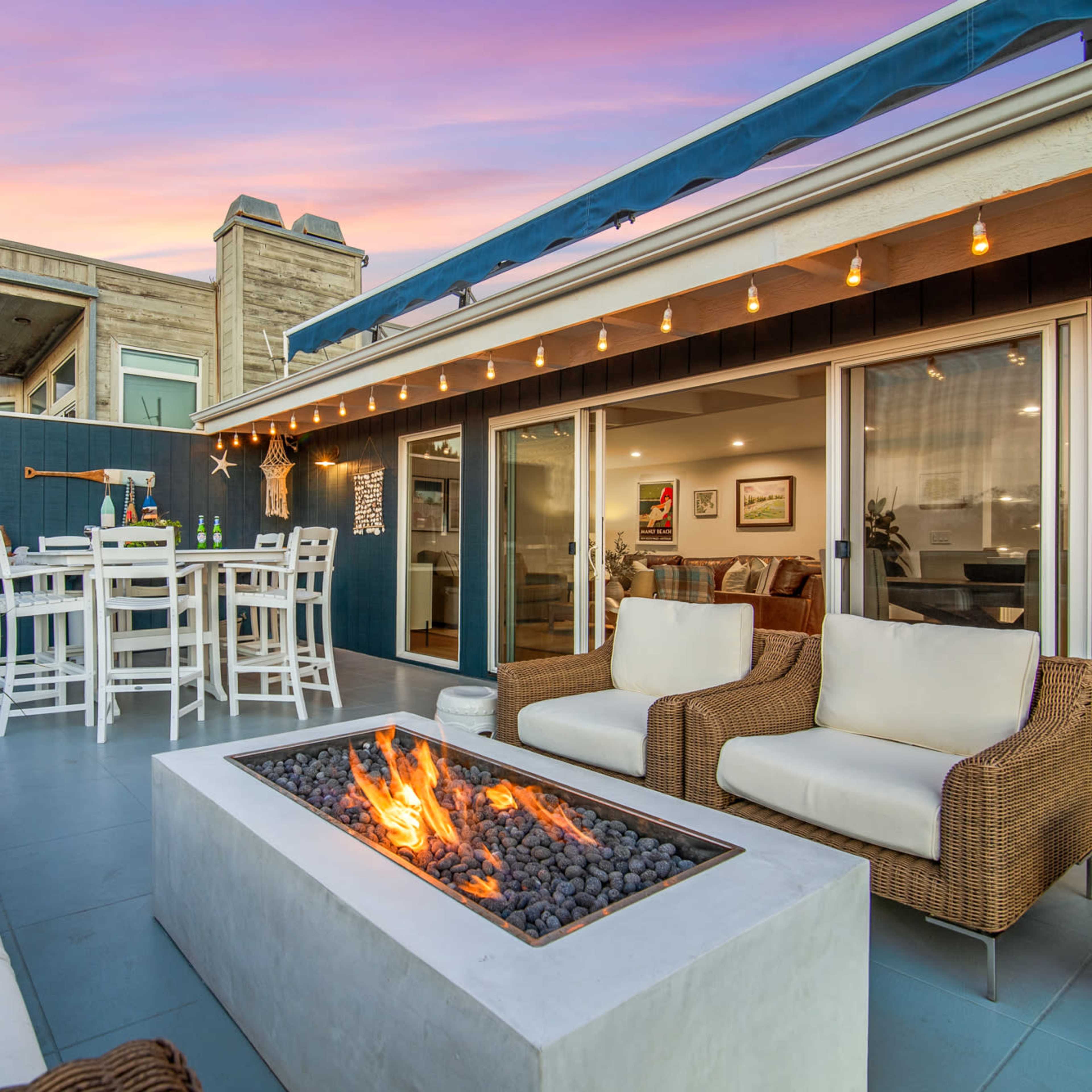 A modern outdoor patio features a fire pit surrounded by seating, with a dining area and a view of a colorful sunset.