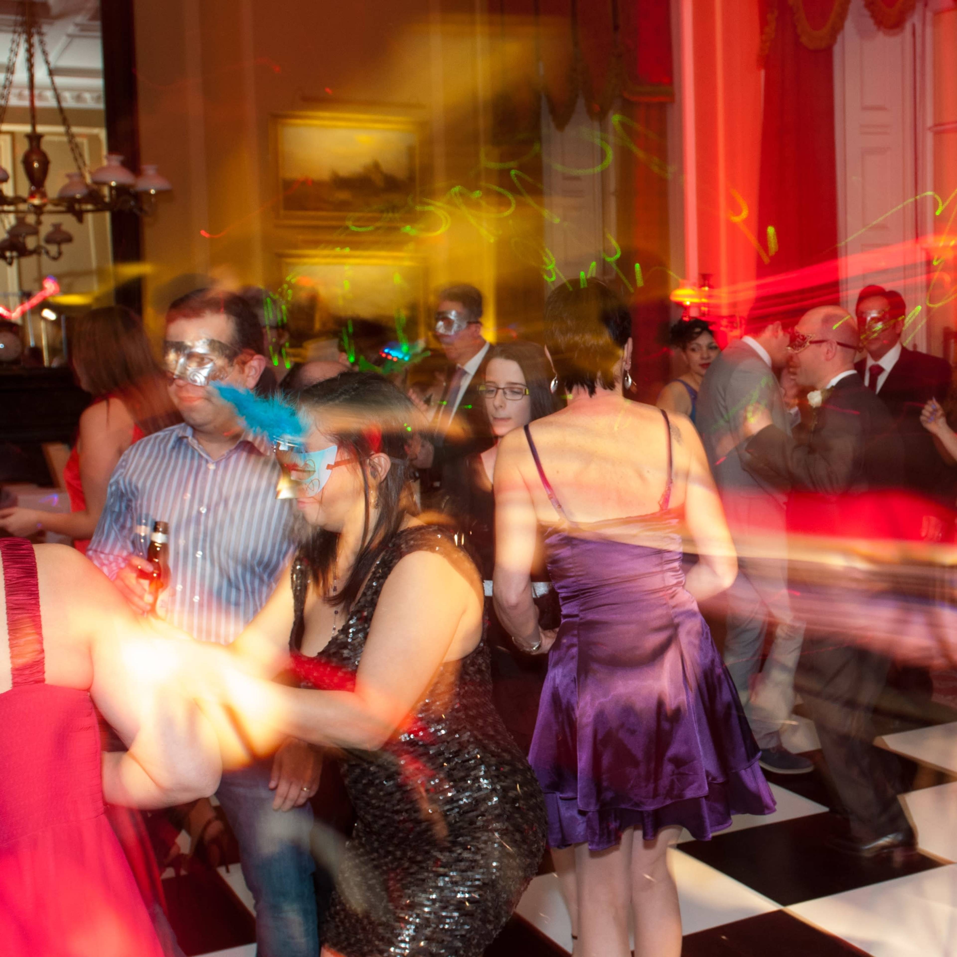 A lively dance party takes place in a elegantly decorated room with guests wearing masks and colorful lights creating streaks in the air.