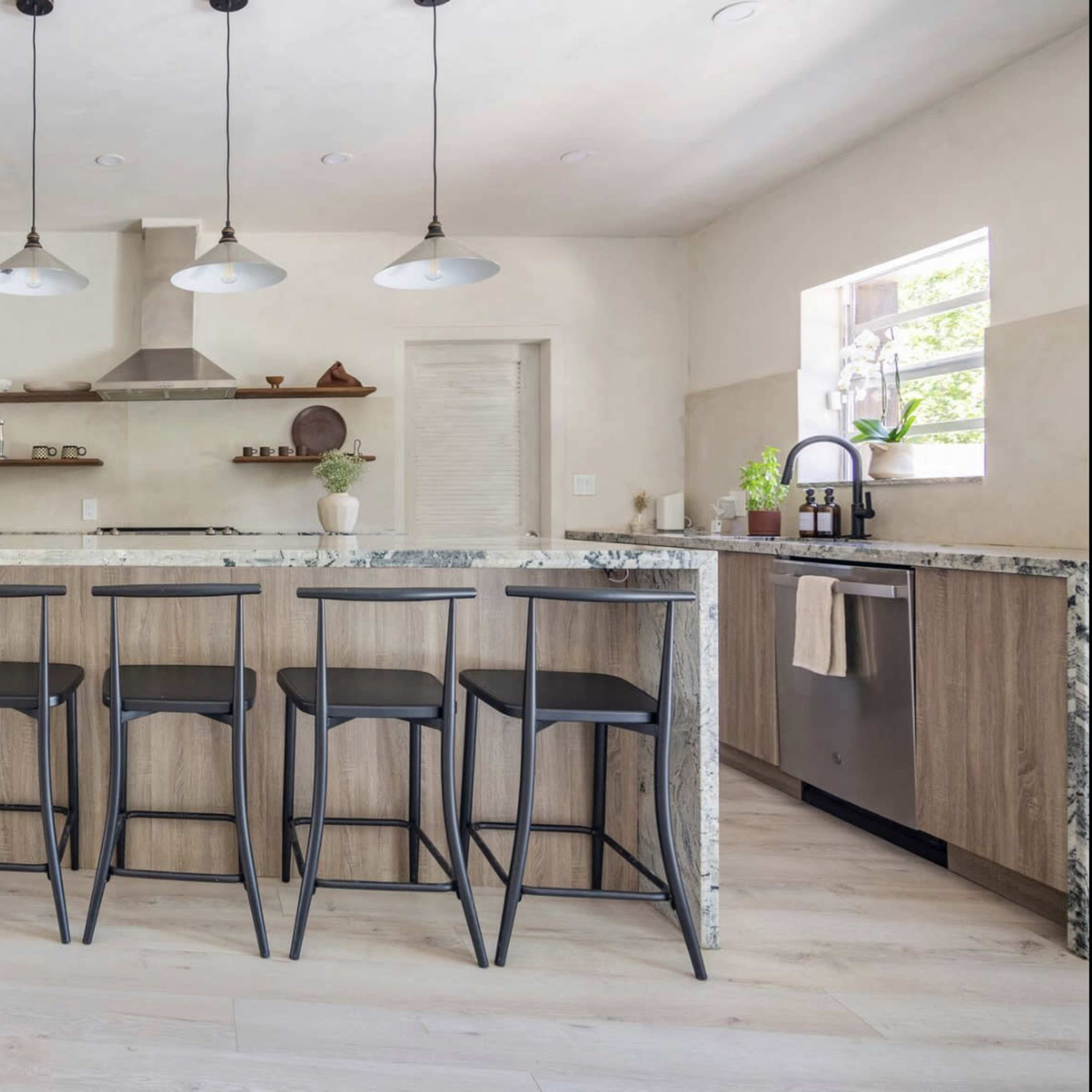 A modern kitchen featuring a large marble-topped island with four black bar stools, stainless steel appliances, and minimalist cabinetry.