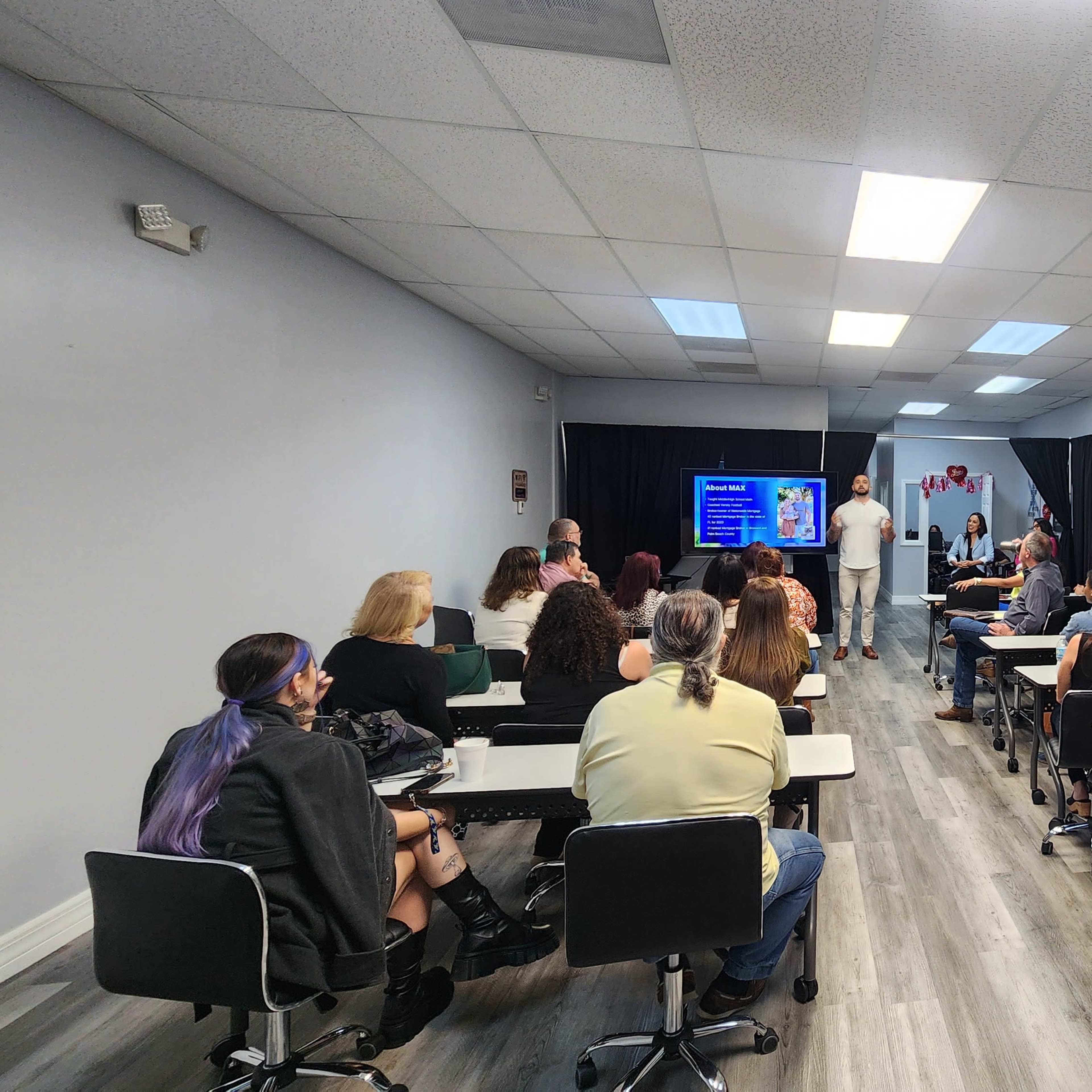 A group of people is seated at desks in a classroom, facing a presenter who stands at the front near a large screen displaying information.