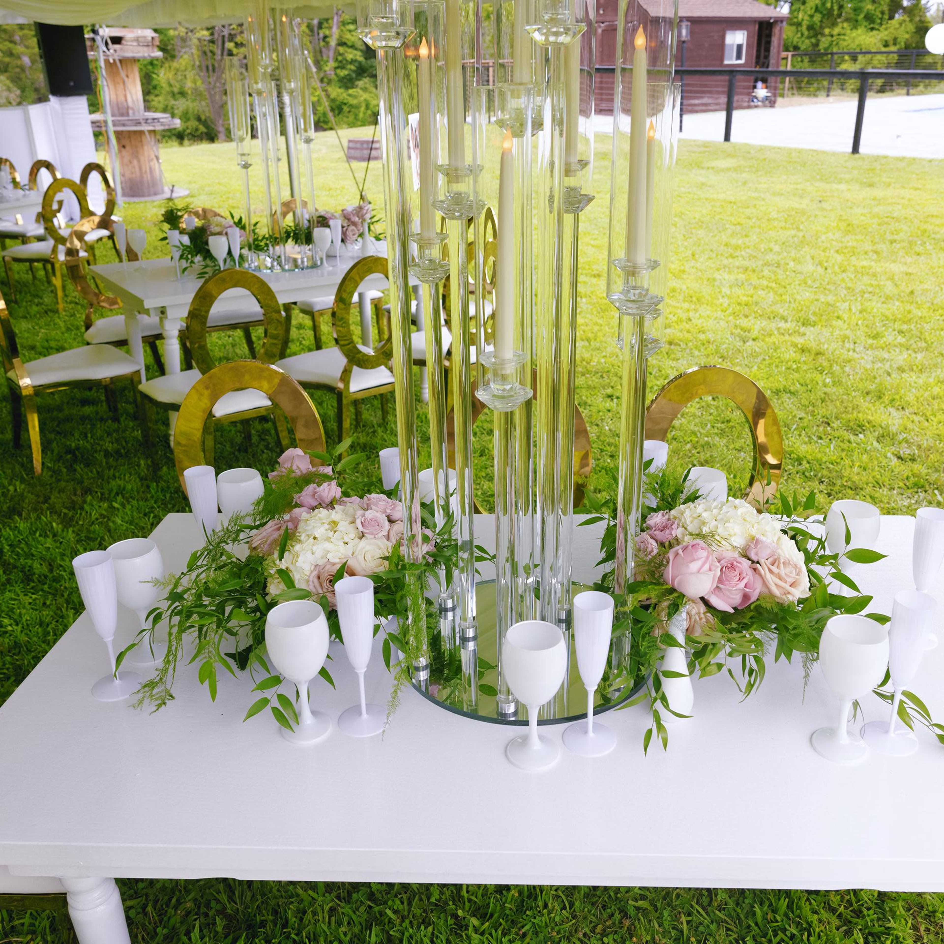 A white dining table adorned with floral arrangements and glass centerpieces is set under a tent on a grassy outdoor venue.