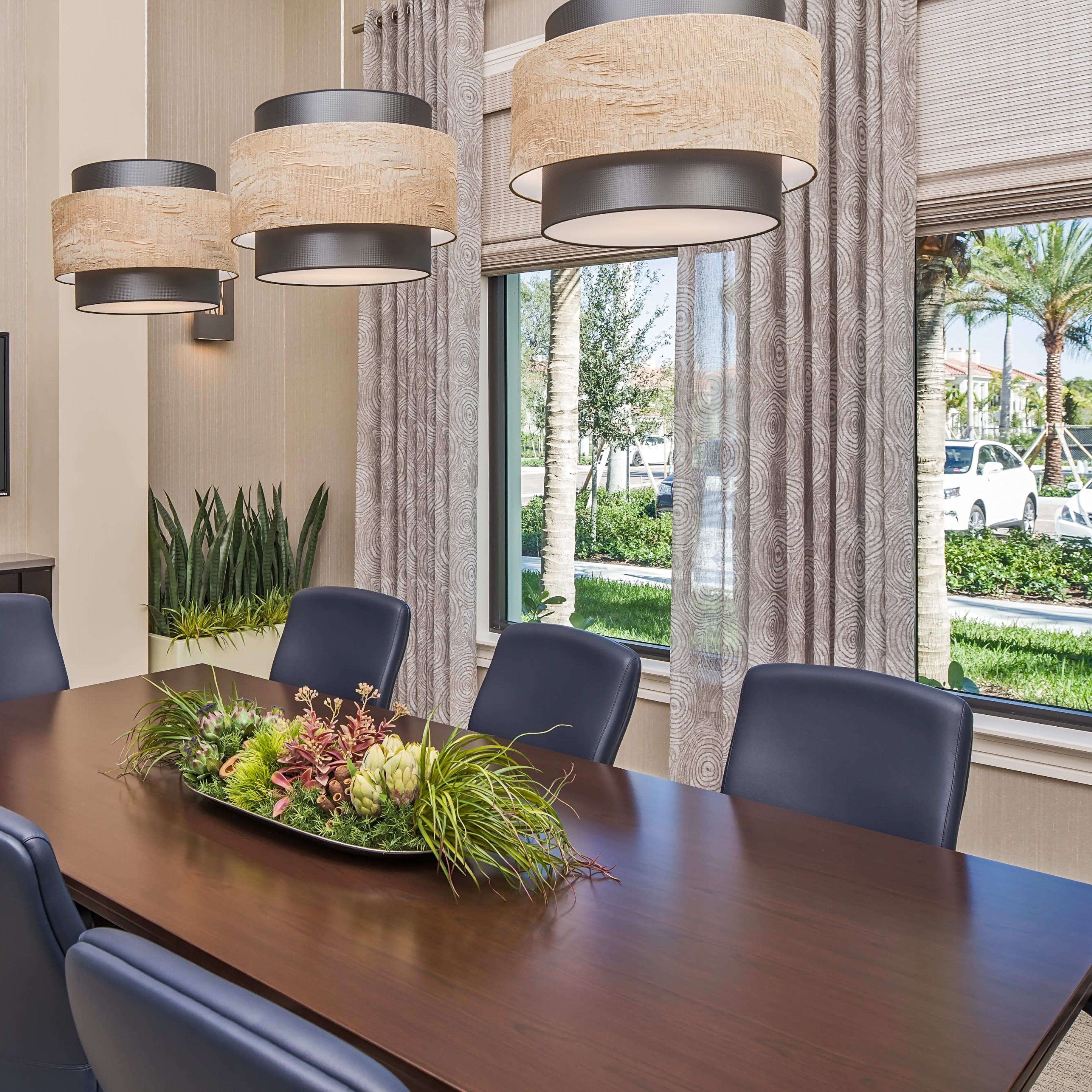 A modern conference room features a long wooden table surrounded by blue chairs, with a TV mounted on the wall and large windows displaying greenery outside.