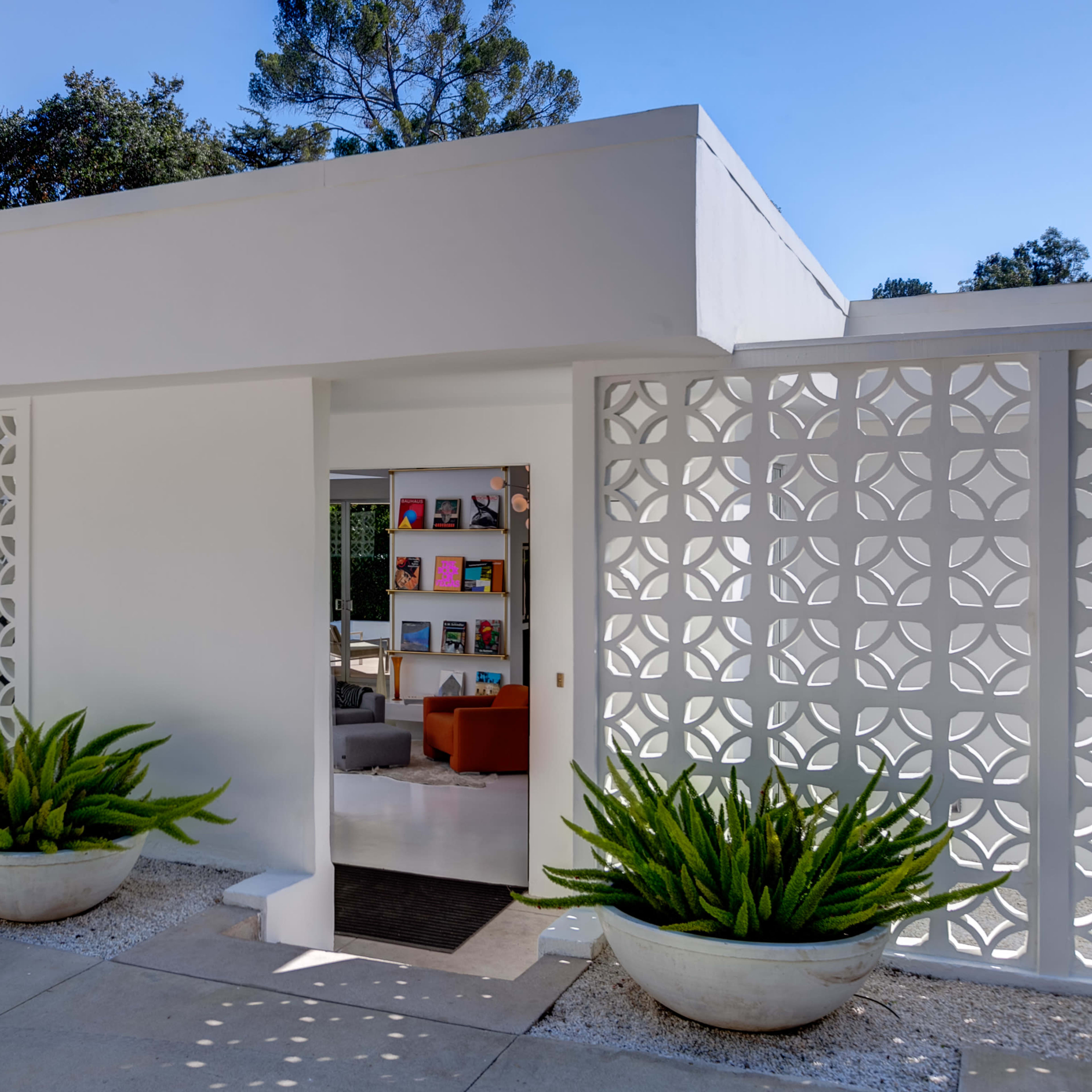 A modern white entrance with decorative lattice walls and potted plants on either side, leading into a brightly lit interior space.