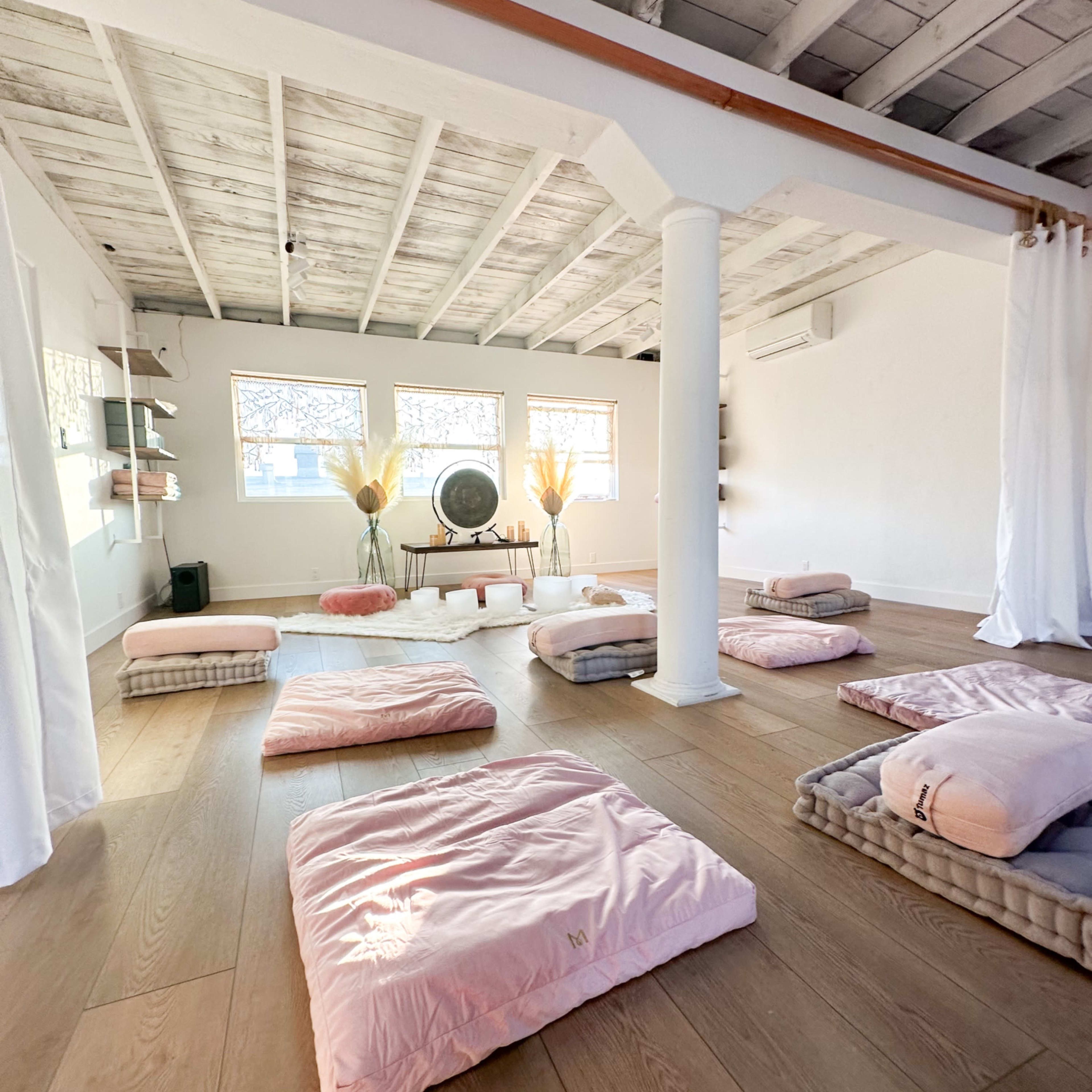 A spacious, minimalist meditation room with several pink mats arranged on a wooden floor, complemented by soft lighting and a large wall fan.