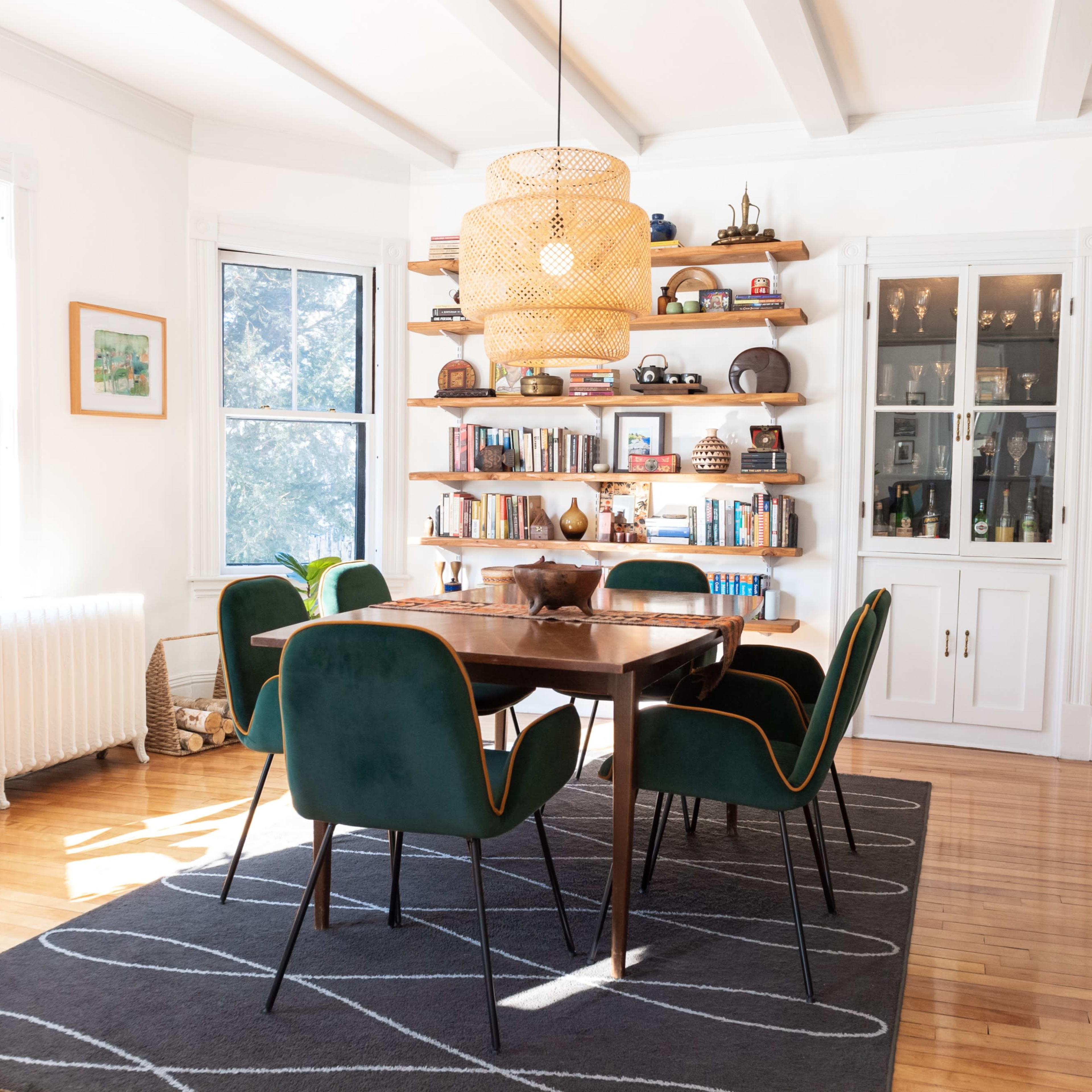 A dining room features a wooden table surrounded by green chairs, with bookshelves and a large light fixture overhead.