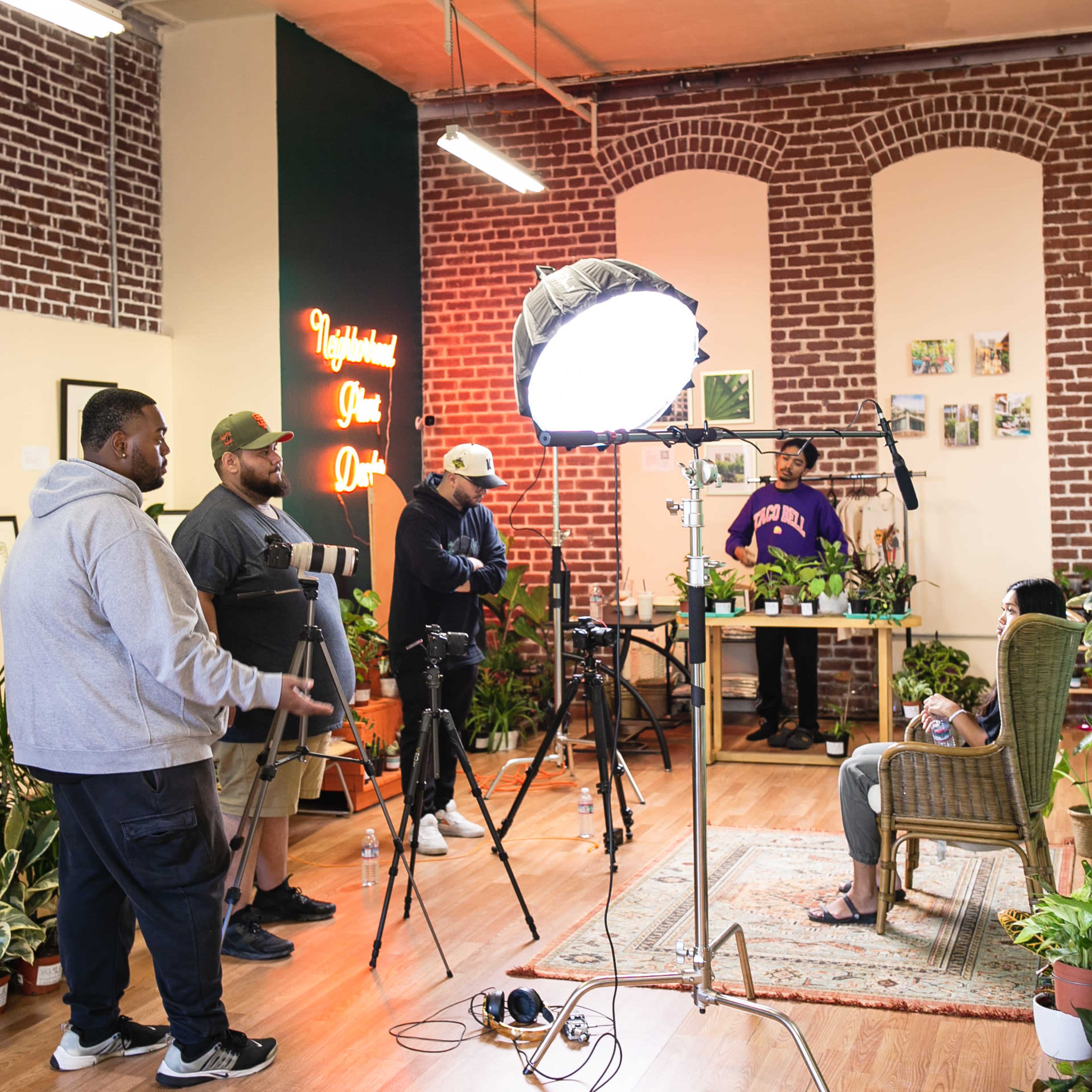 A group of four individuals sets up video equipment in a room filled with plants and brick walls.