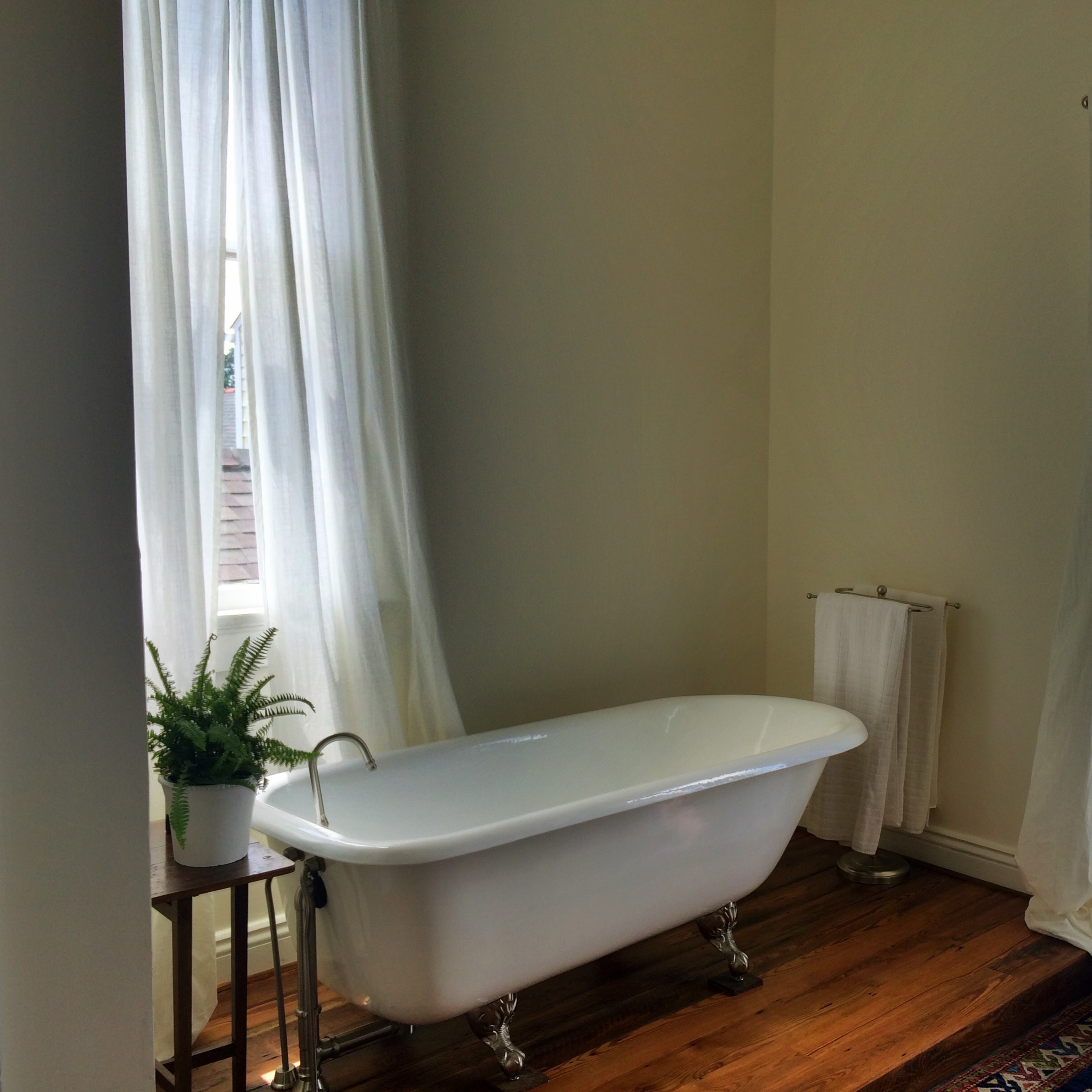 A bright bathroom featuring a freestanding white bathtub next to a window, with light curtains and wooden flooring.