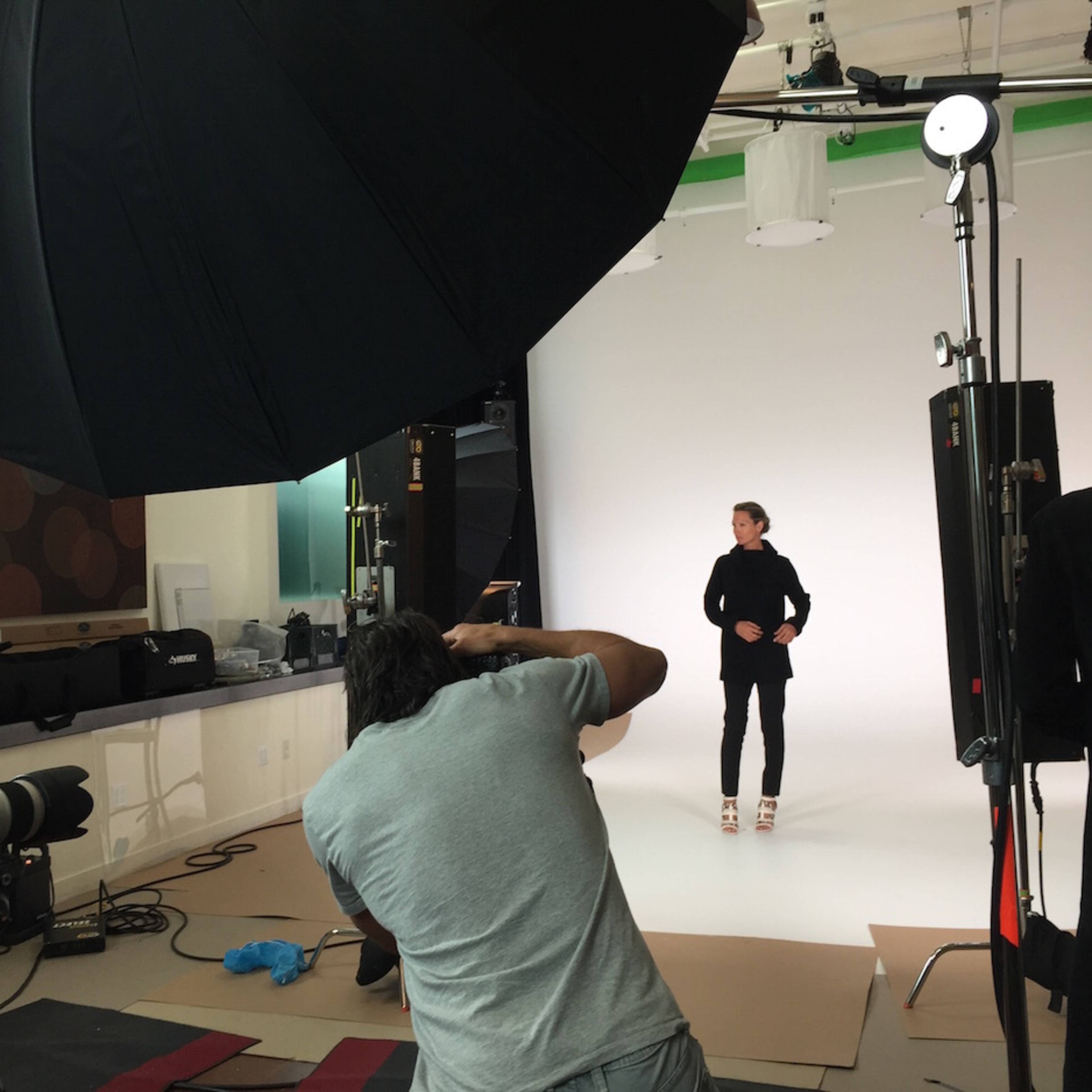 A photographer captures a model in a photo studio with a large softbox light overhead and equipment arranged around a seamless white backdrop.