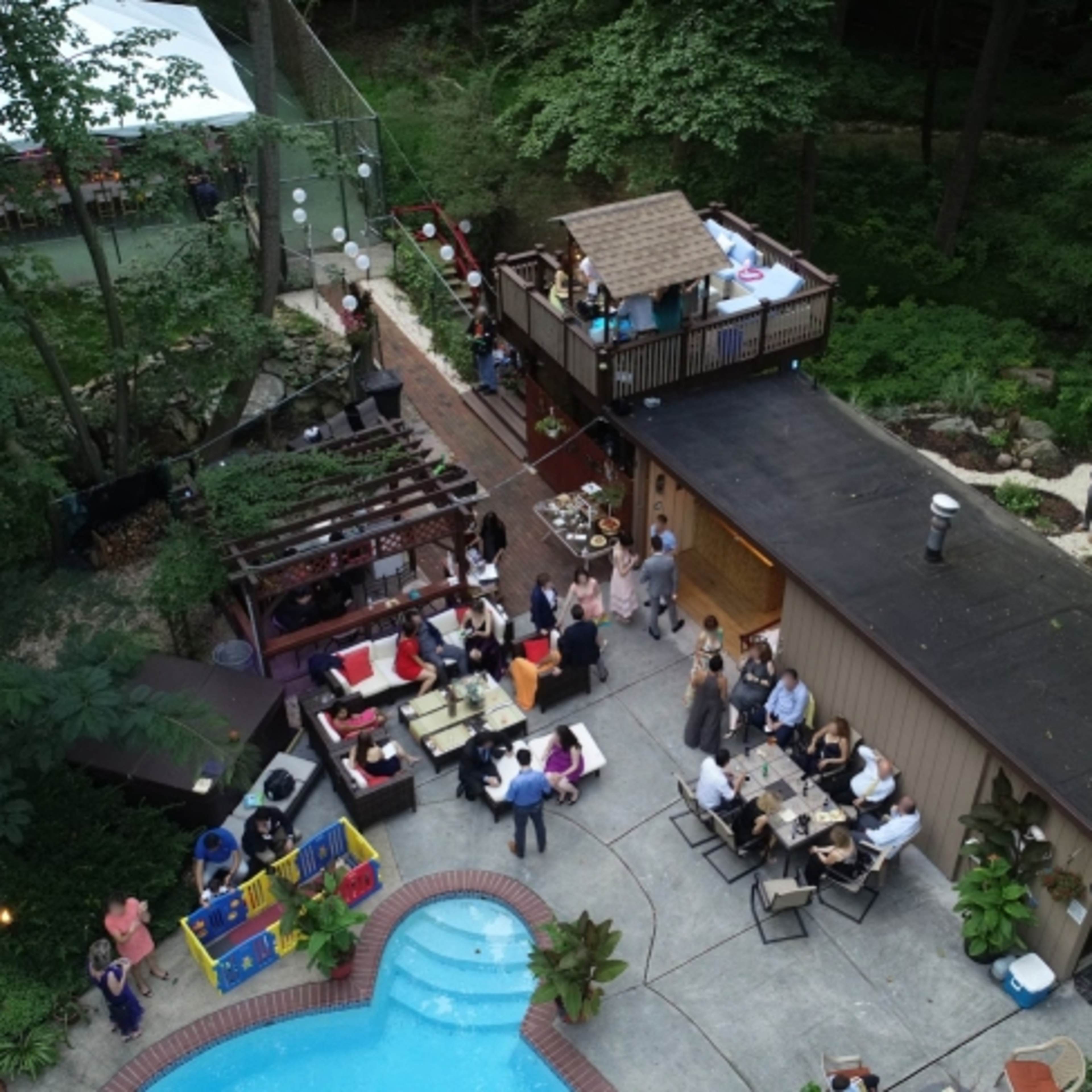 An outdoor gathering takes place around a pool, with guests socializing near a house and a separate deck area surrounded by trees.