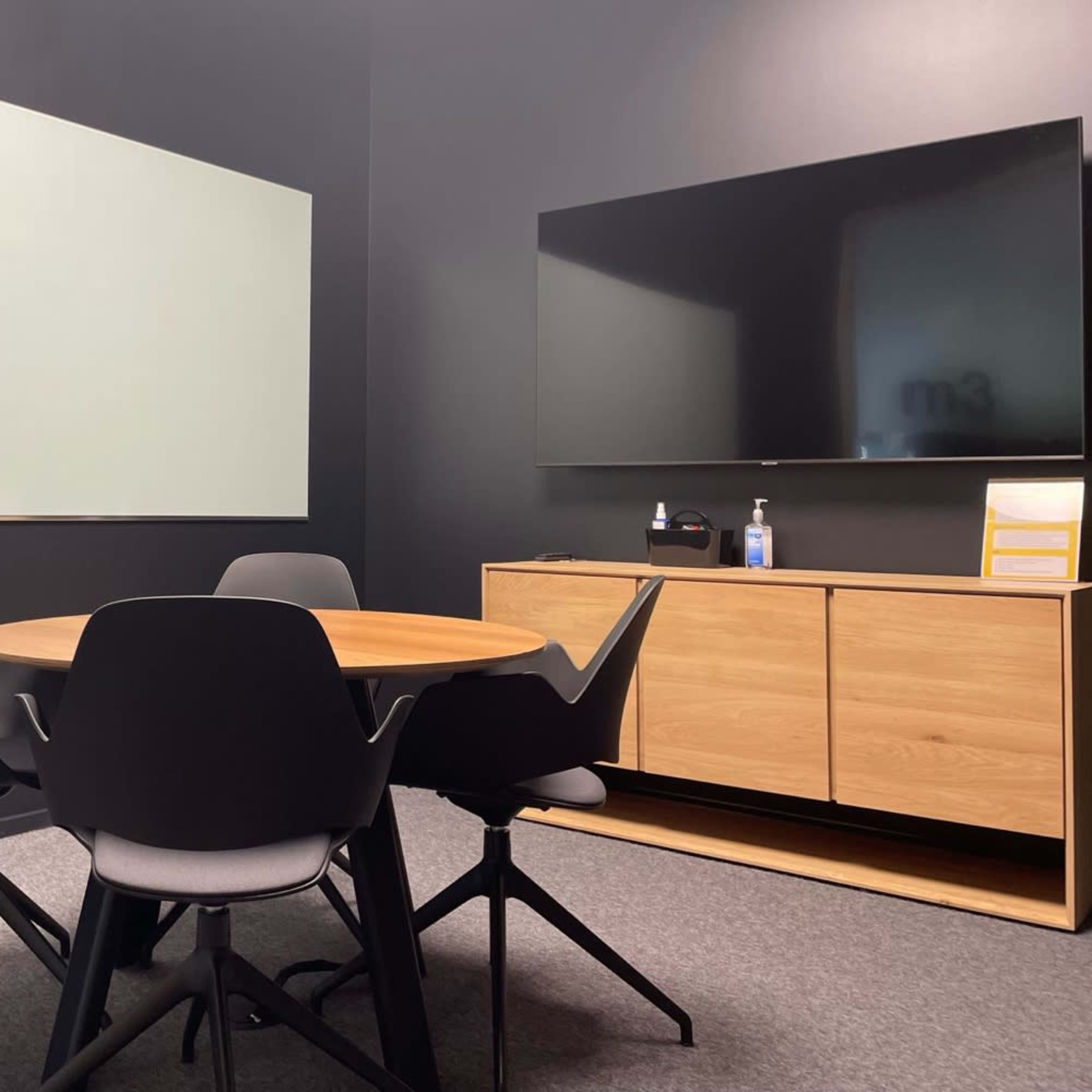A small conference room featuring a round wooden table surrounded by black chairs, a whiteboard, and a wall-mounted television.