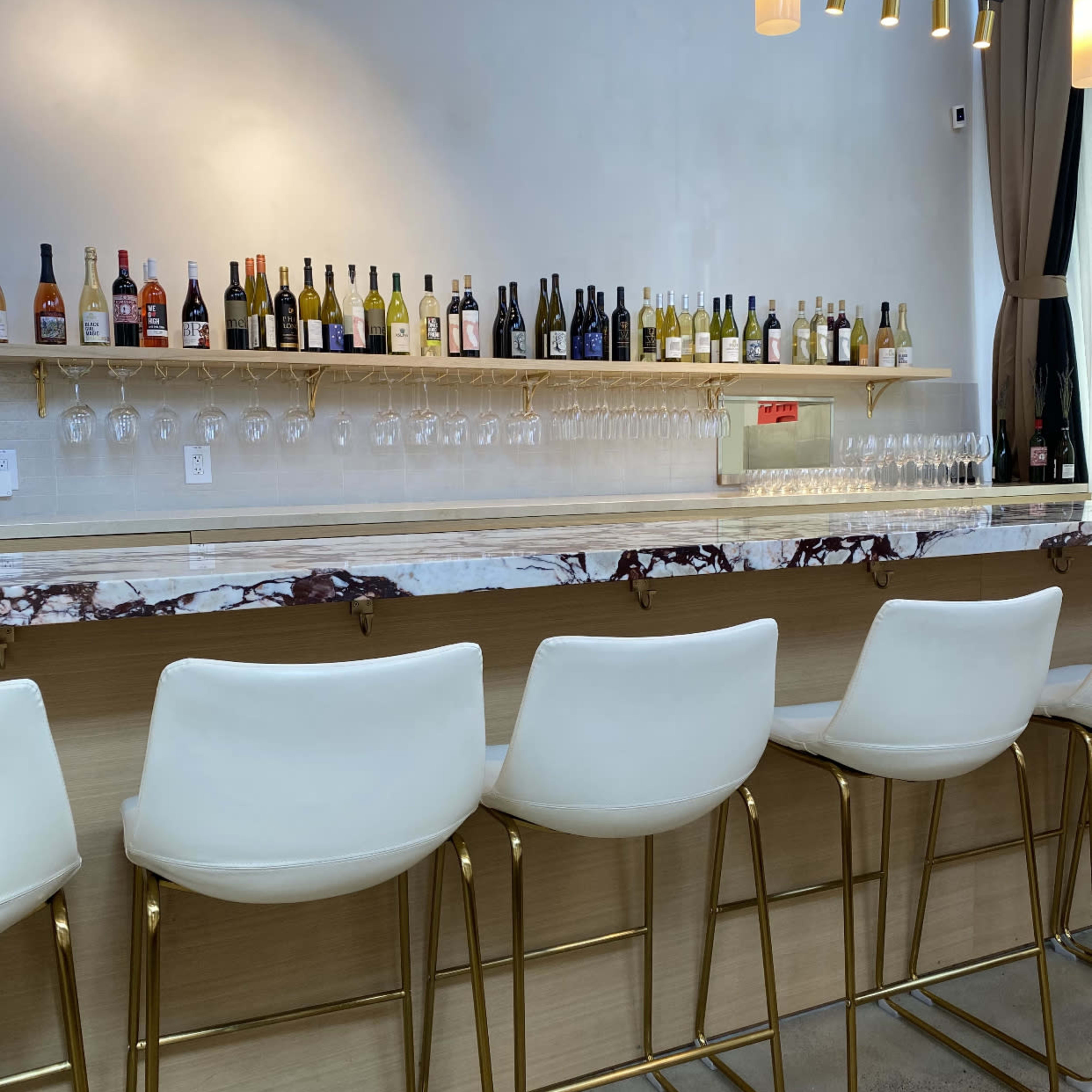 A modern bar area features a marble countertop, white stools with gold legs, and a shelf lined with various bottles of liquor and wine.