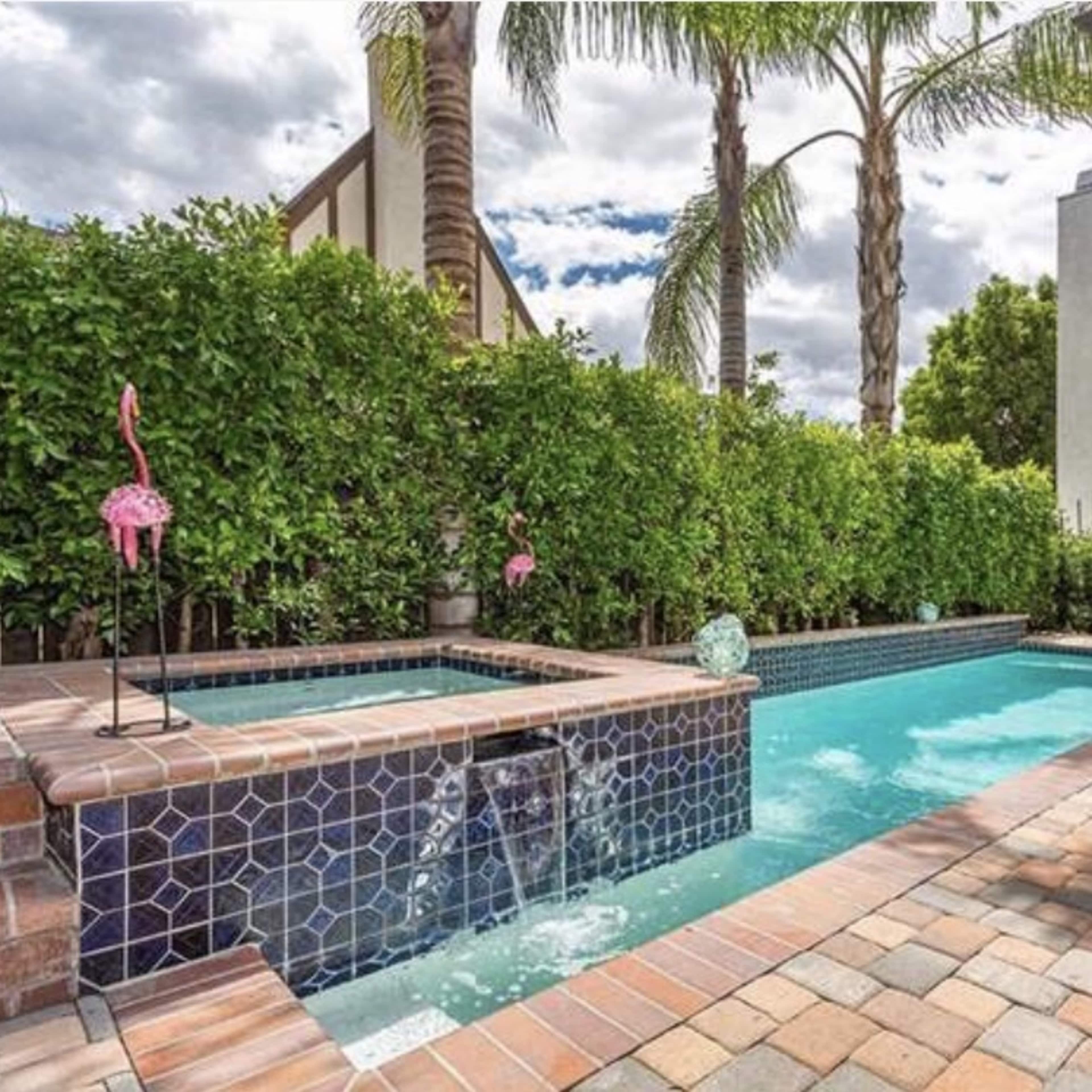 A pool area with a spa, surrounded by greenery and palm trees, featuring decorative flamingos and brick paving.