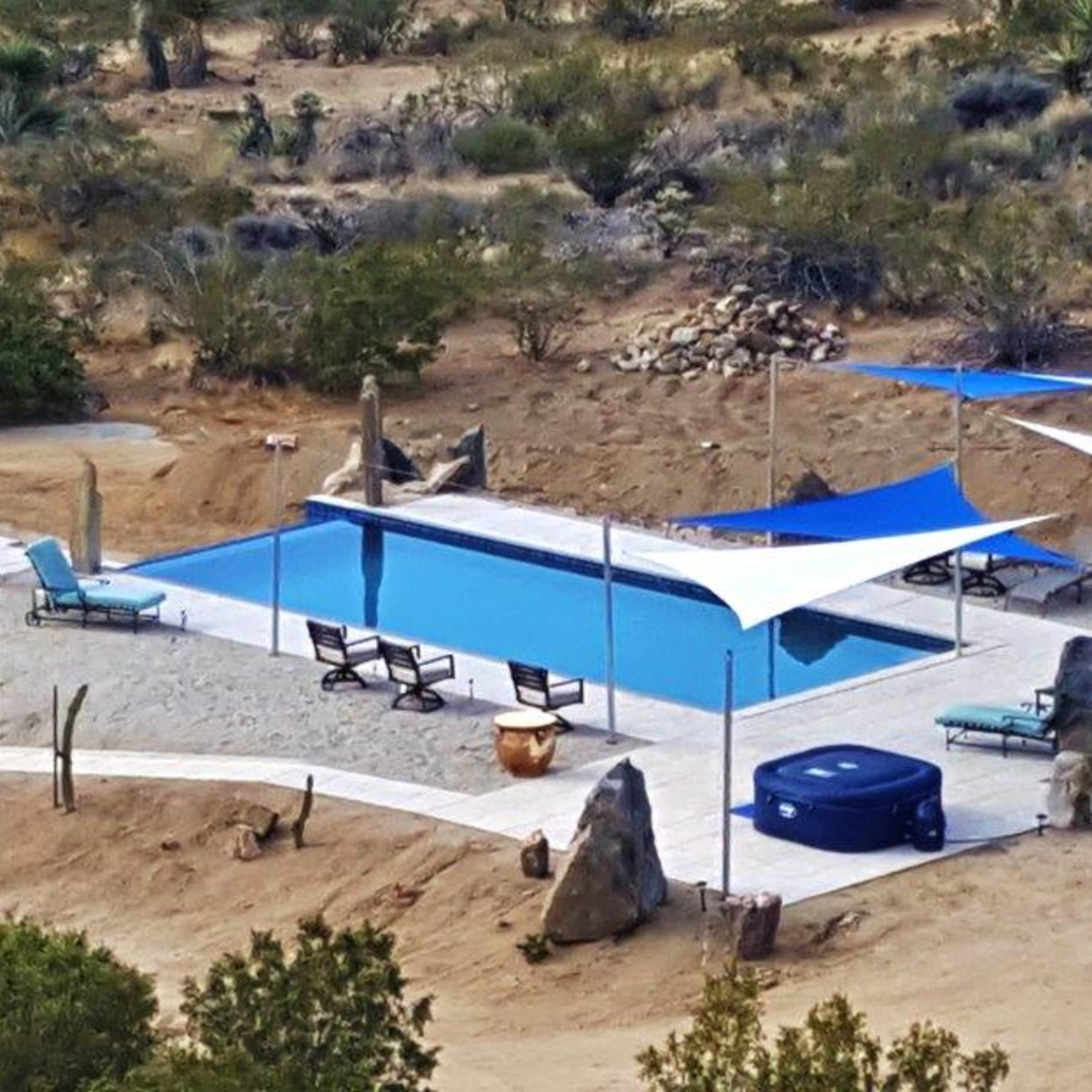 A rectangular swimming pool surrounded by lounge chairs and shaded by white canopies in a desert landscape.