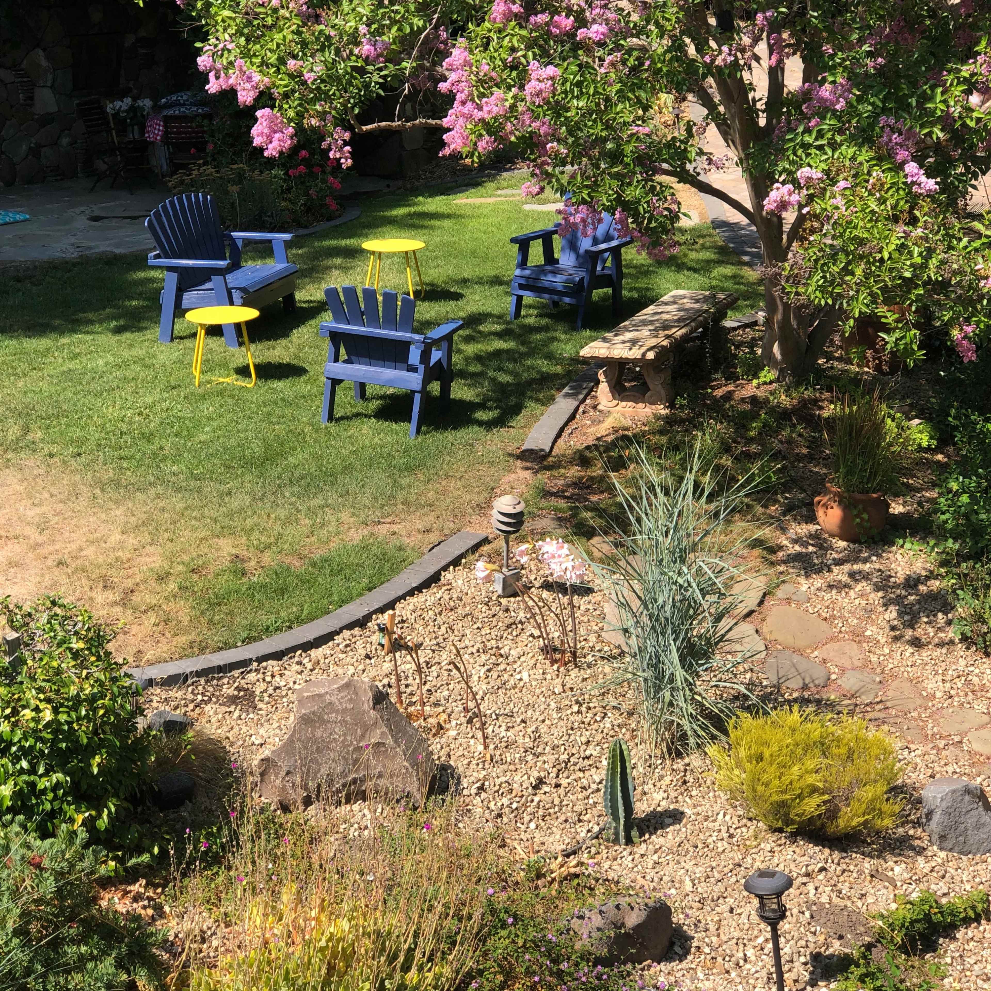 A garden with purple flowering trees, green grass, and blue and yellow chairs arranged around a stone path.