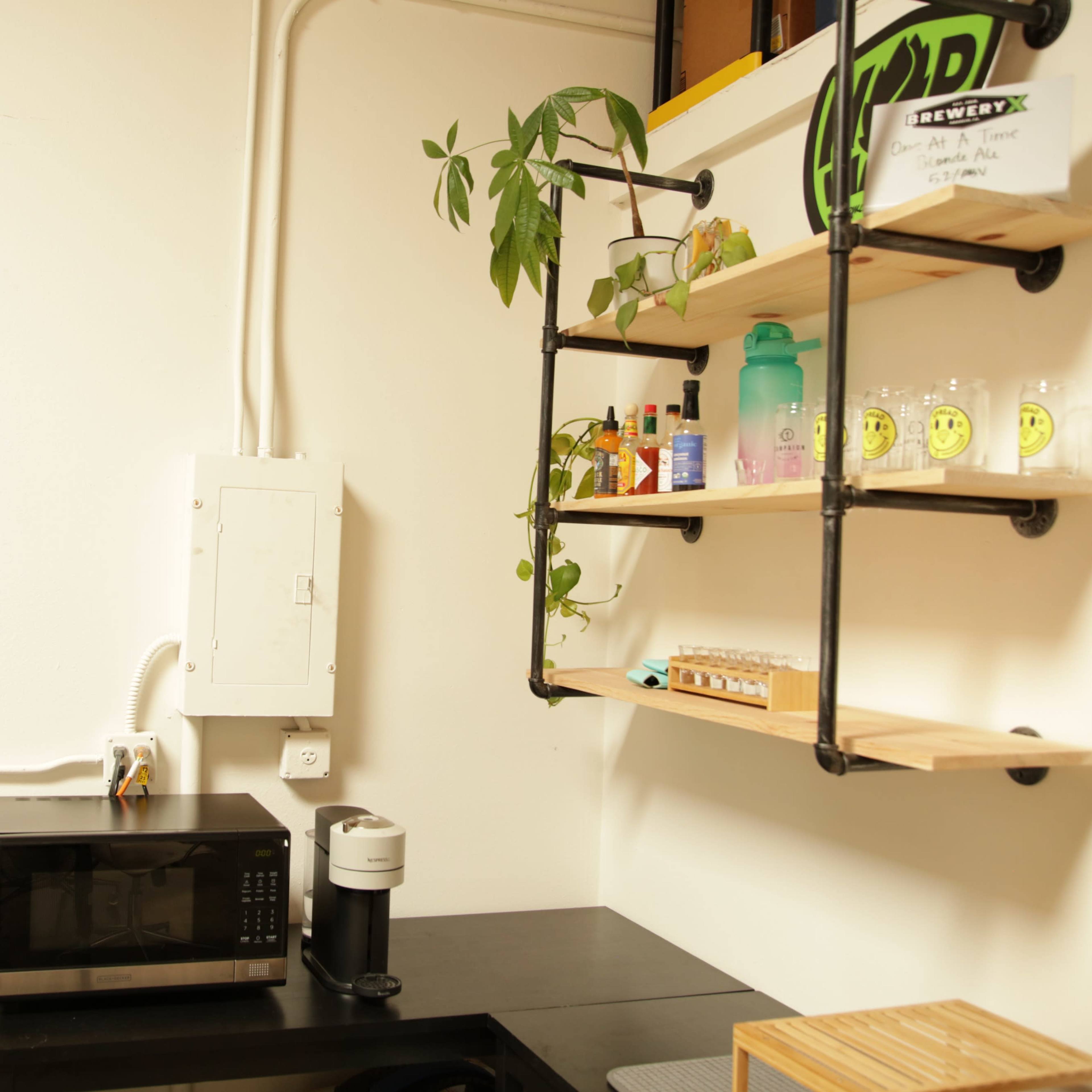 A small kitchen area with a microwave, coffee maker, and shelves made of wood and metal mounted on a white wall, displaying various bottles and plants.