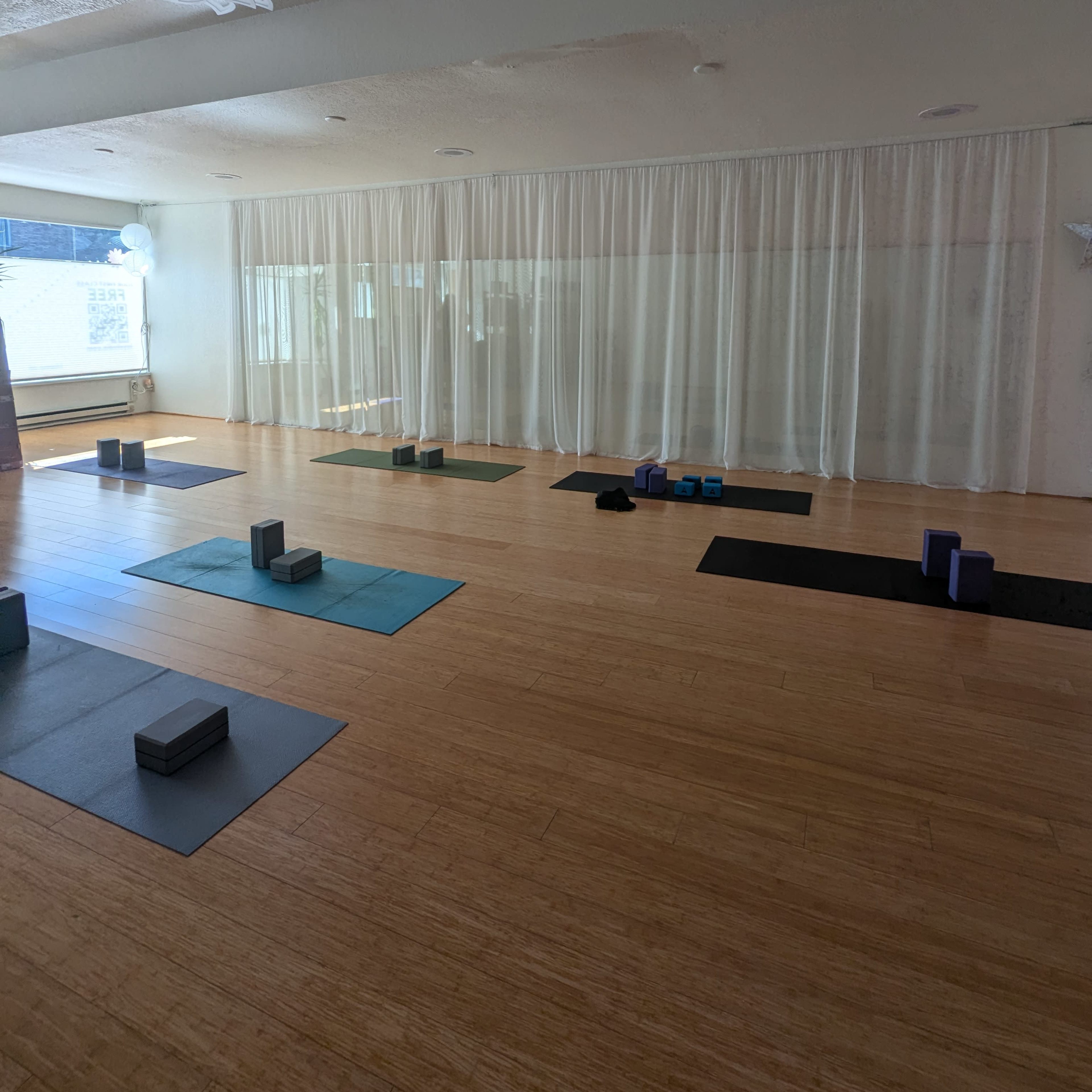A spacious yoga studio features bamboo flooring, exercise mats, and props arranged neatly, with sheer curtains allowing natural light to filter in.