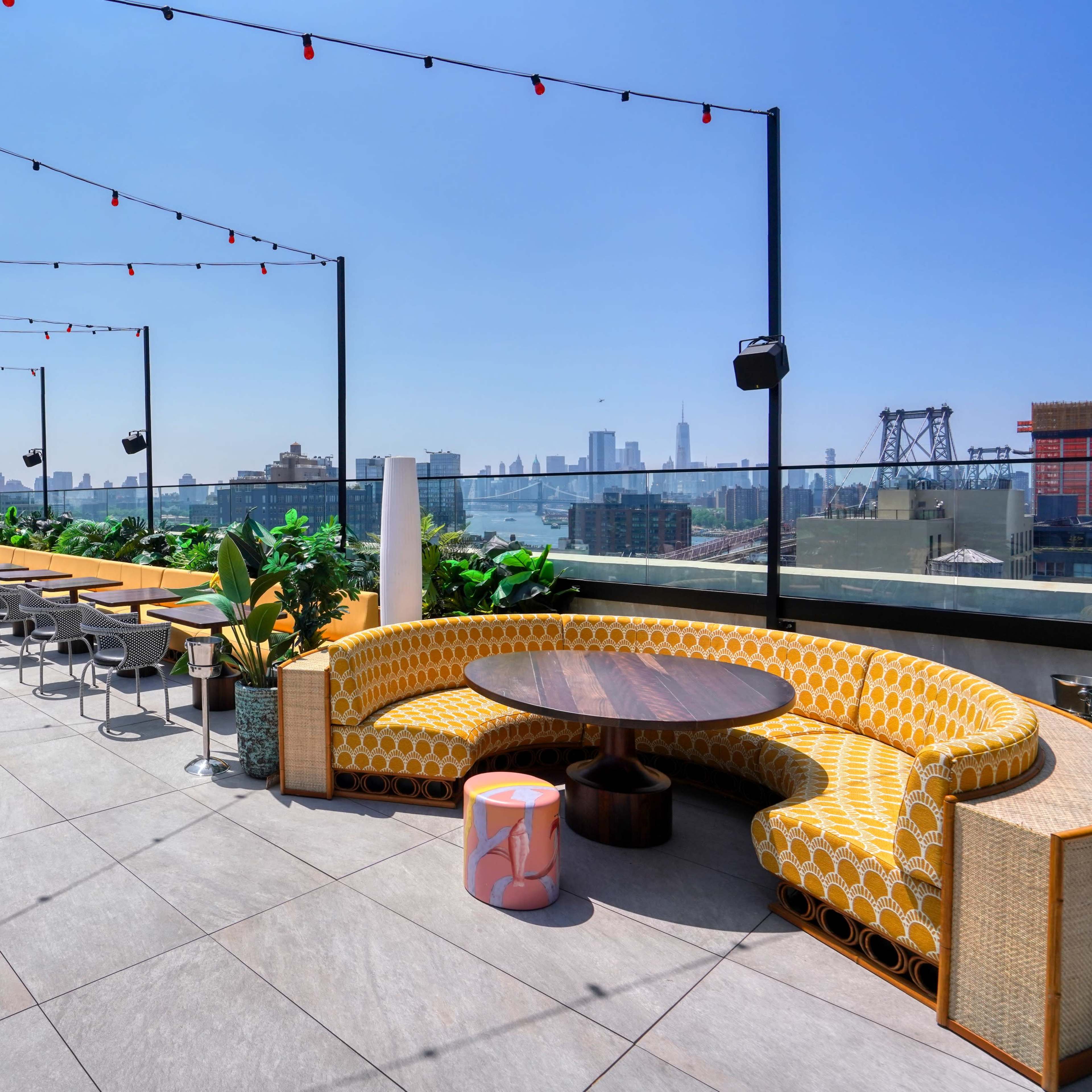 A rooftop terrace features circular and rectangular seating arrangements, tables, and decorative plants with a city skyline in the background.