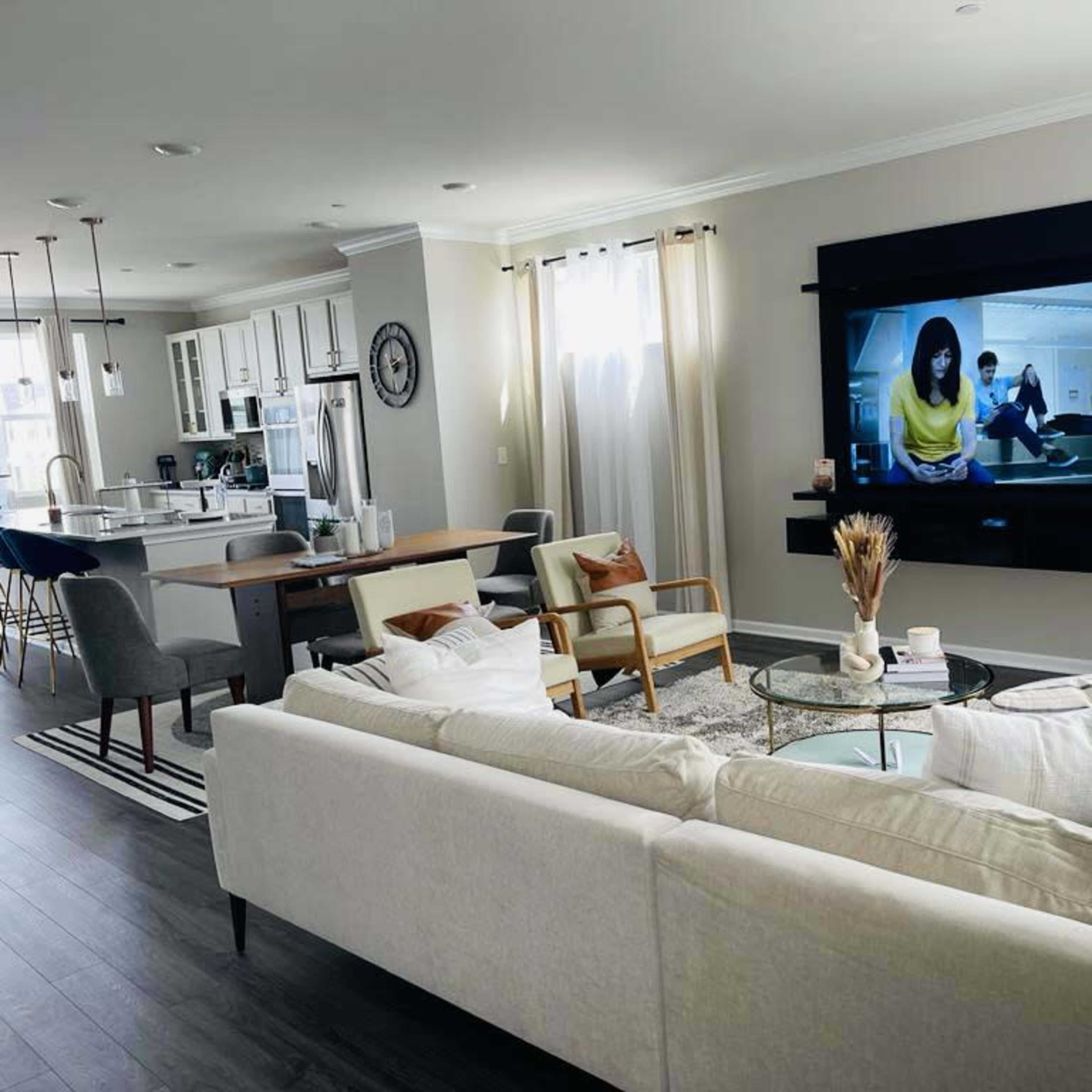 A modern living room features a large sectional sofa facing a mounted television, with a dining area and kitchen visible in the background.