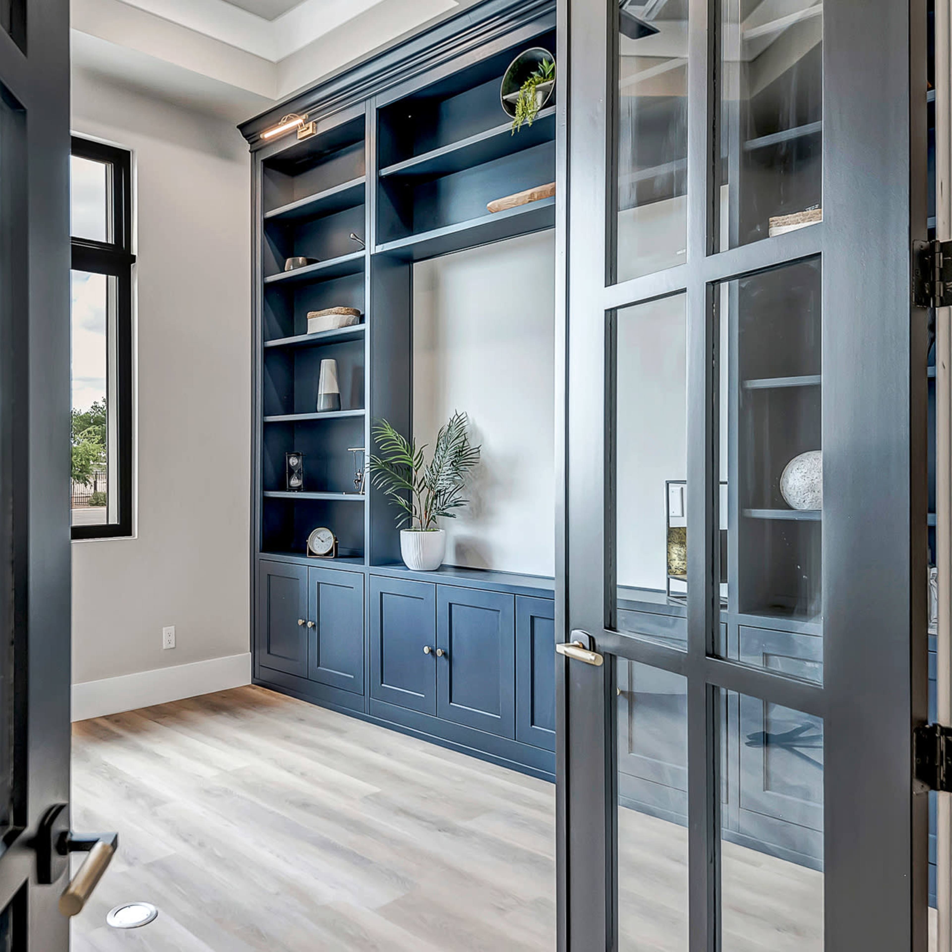 A modern home office features a dark blue built-in bookshelf surrounded by large windows, with double glass doors opening into the space.