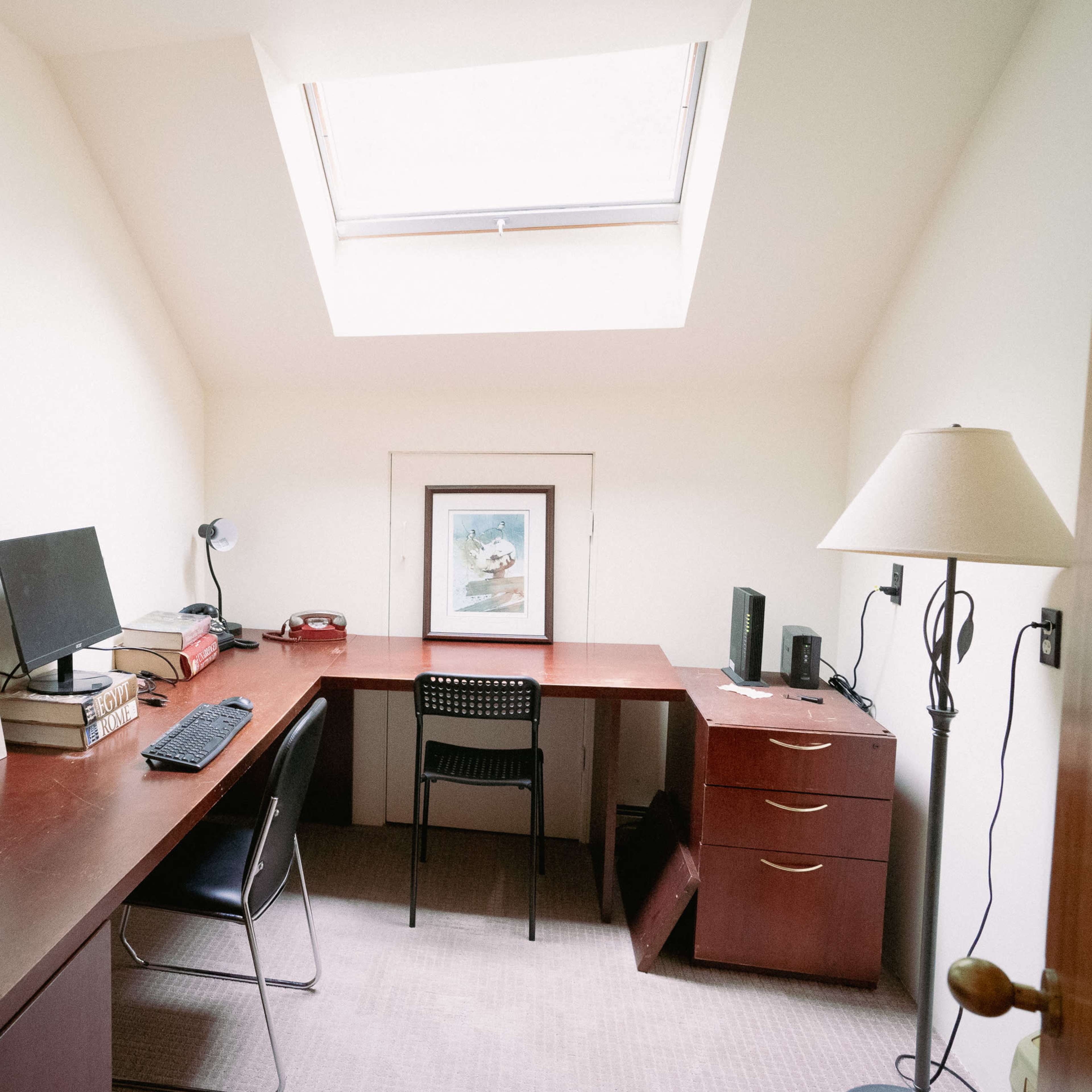 A small, minimalist office features a desk with a computer, books stacked on one side, and a lamp beside a filing cabinet.