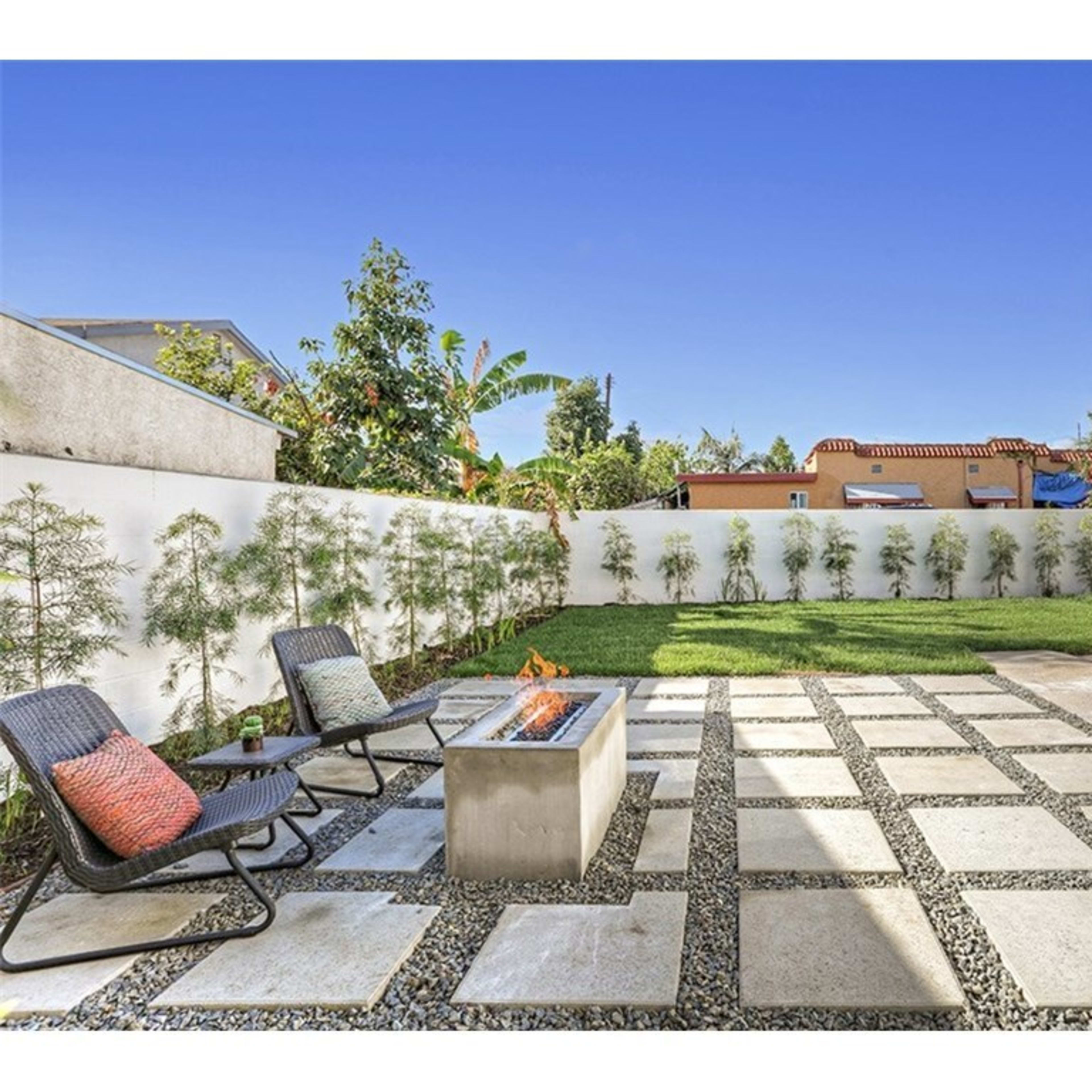 A backyard with a stone patio, a fire pit surrounded by chairs, and a green lawn enclosed by white walls and vegetation.