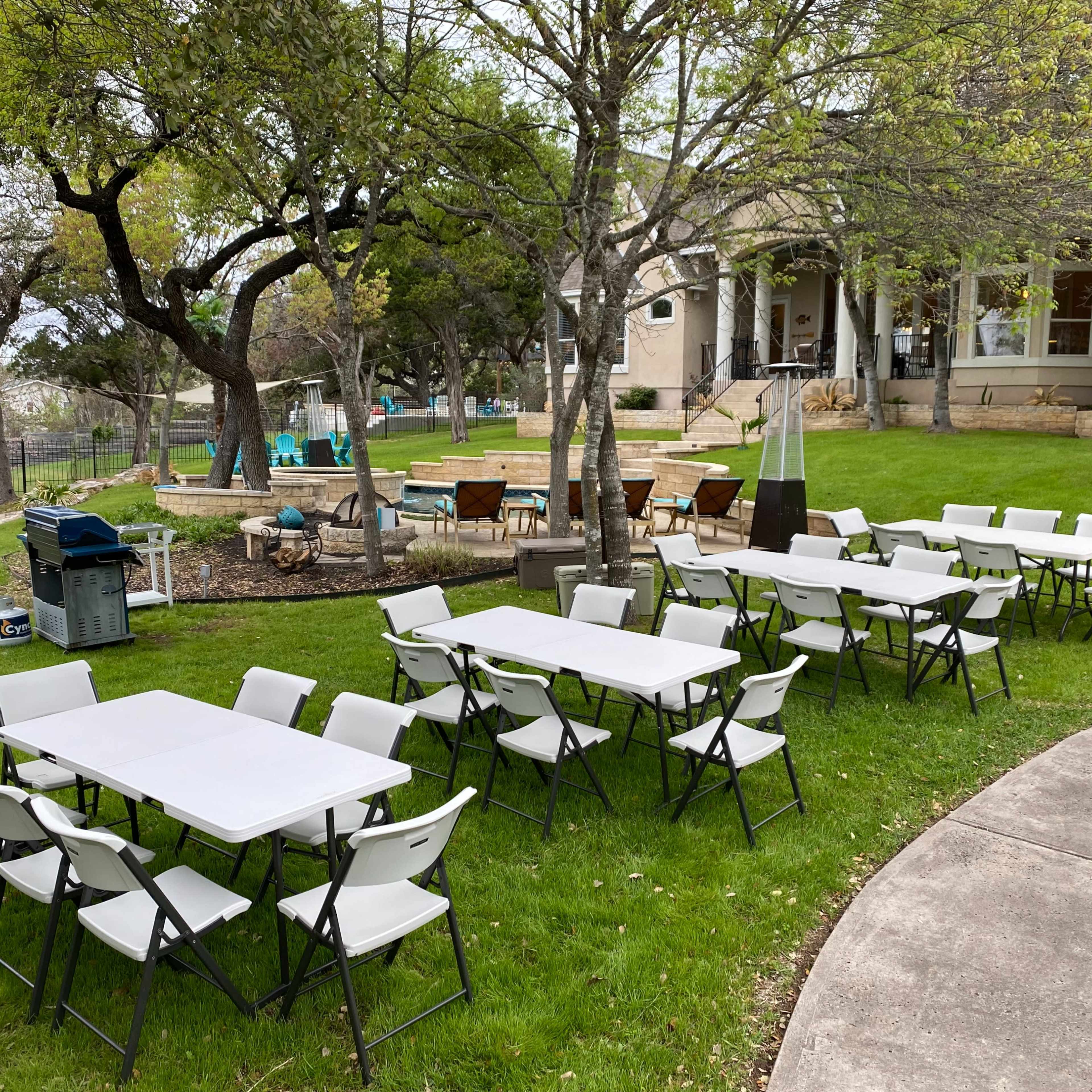 A set of white folding tables and chairs arranged on a grassy area near a house, with trees and a fire pit visible in the background.