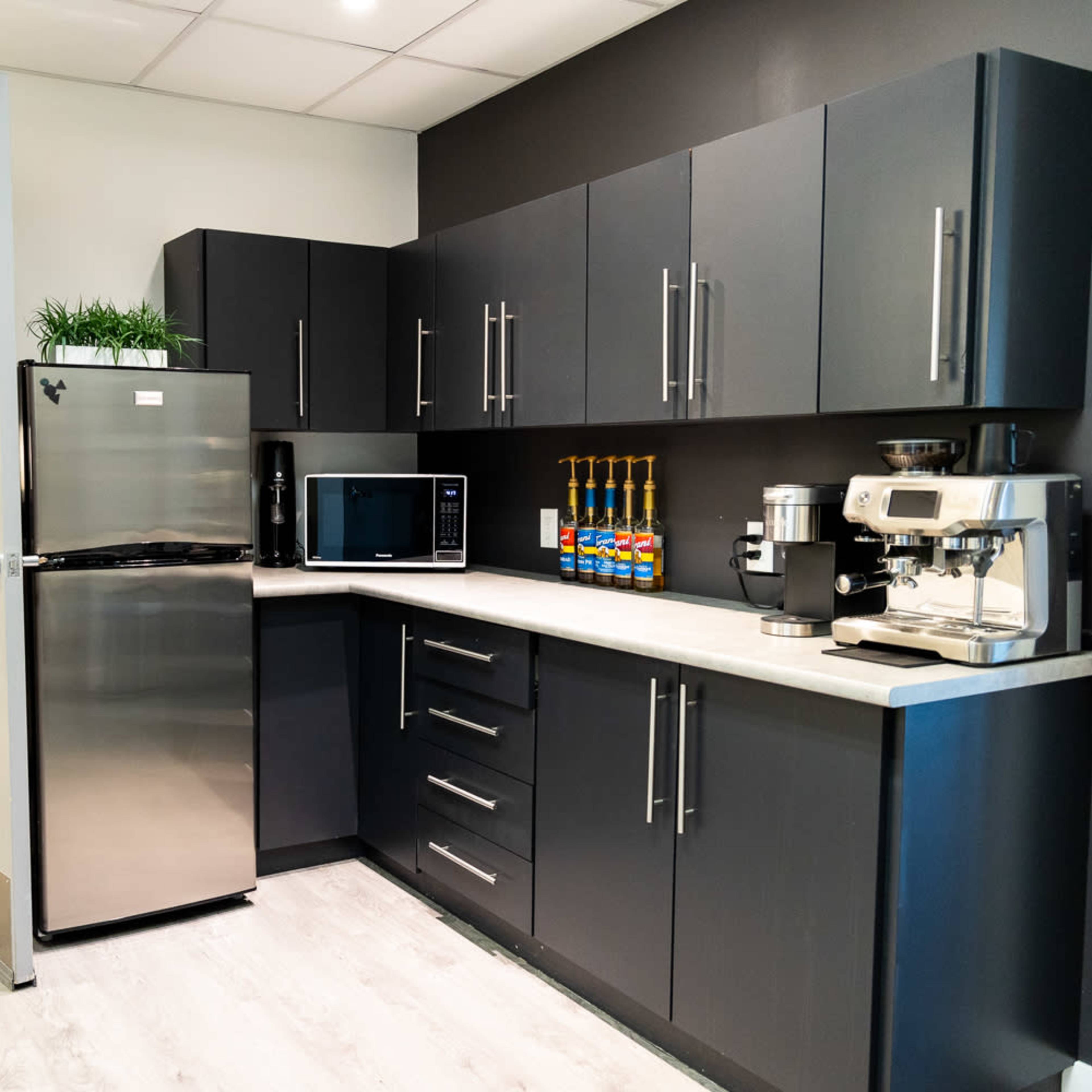 A modern kitchen with dark cabinetry, a stainless steel refrigerator, a microwave, and a coffee machine, alongside a countertop and a door leading to another room.