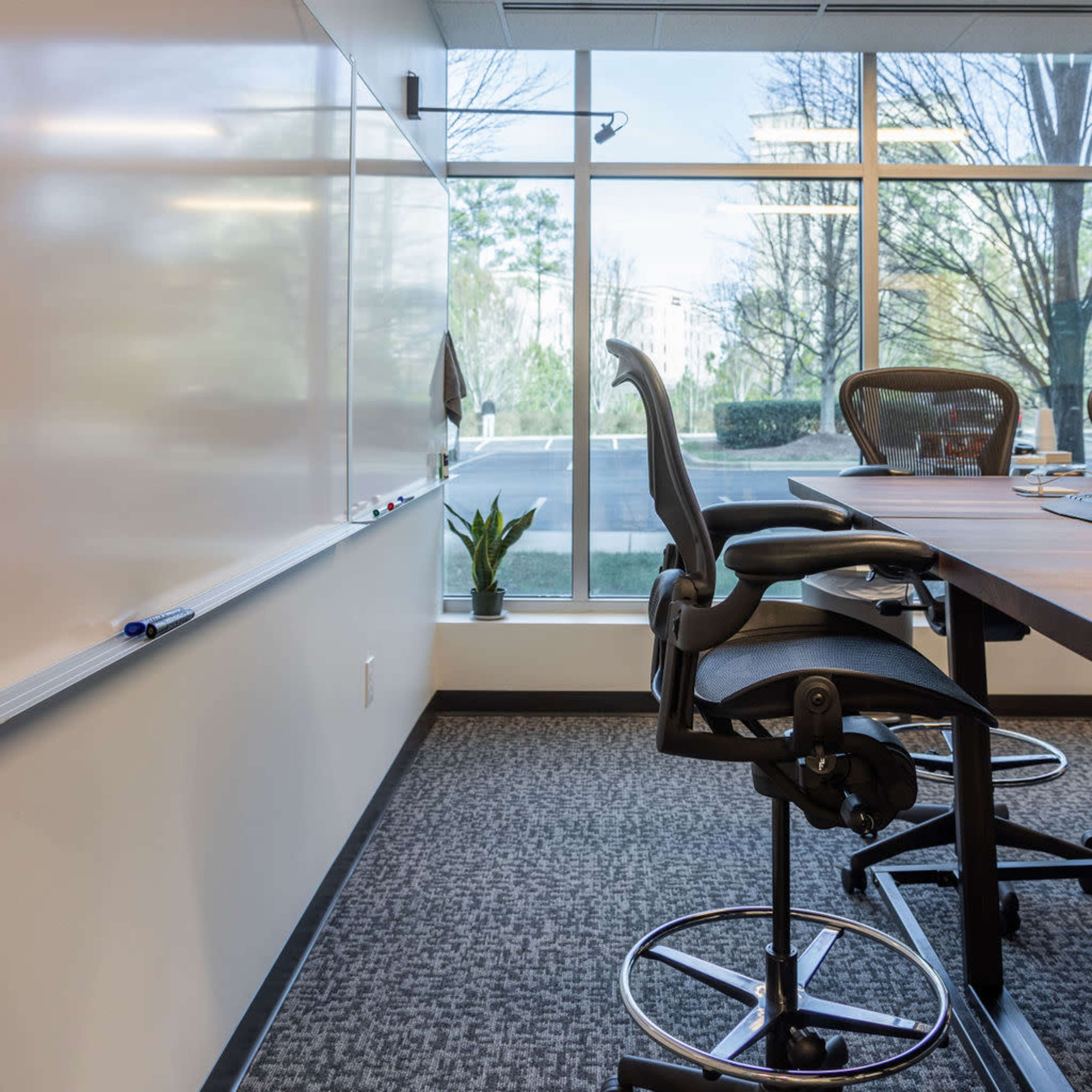 A conference room with a whiteboard, tall chairs, and large windows showcasing greenery outside.