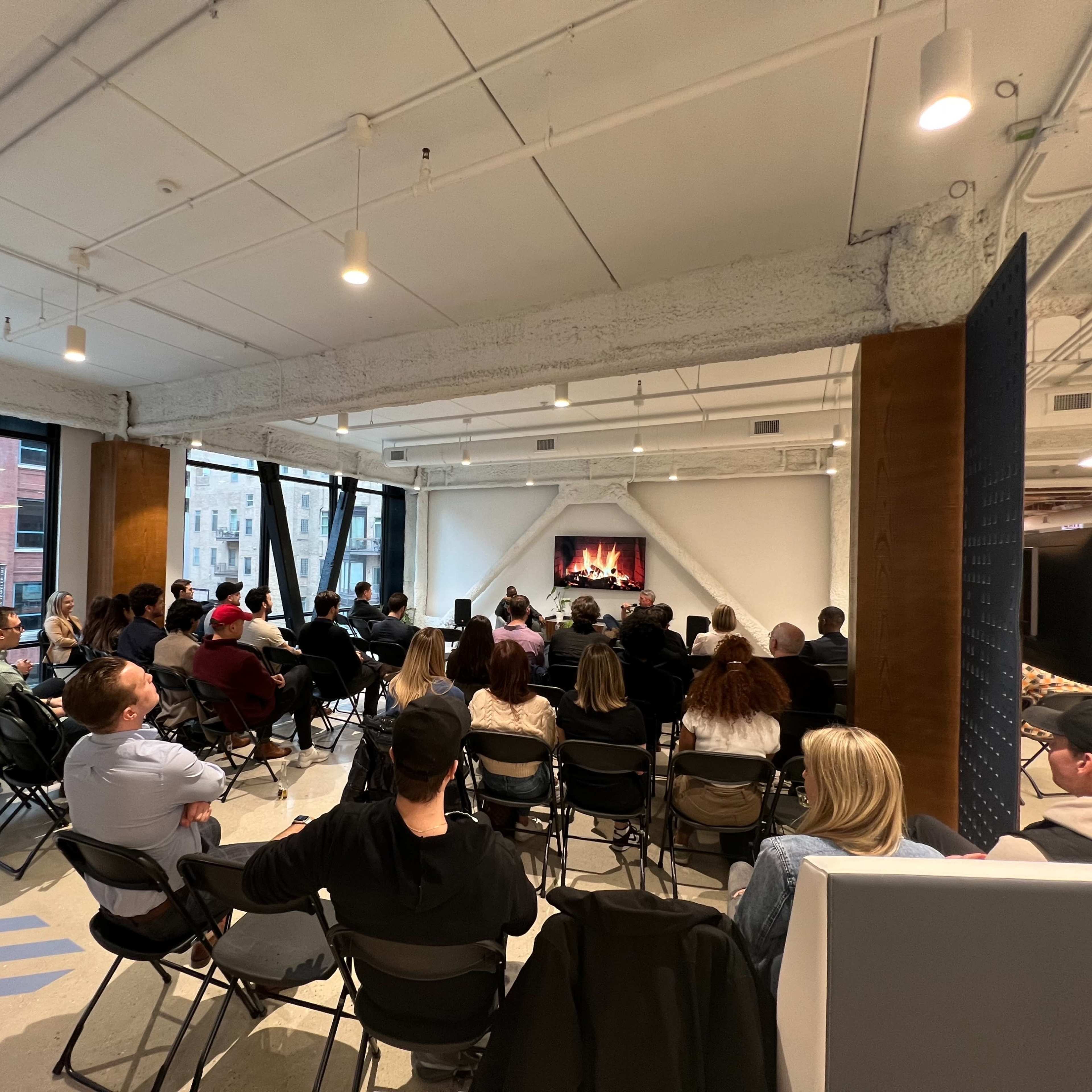 A large audience sits in chairs facing a speaker in a modern indoor space with large windows and a digital fireplace display.