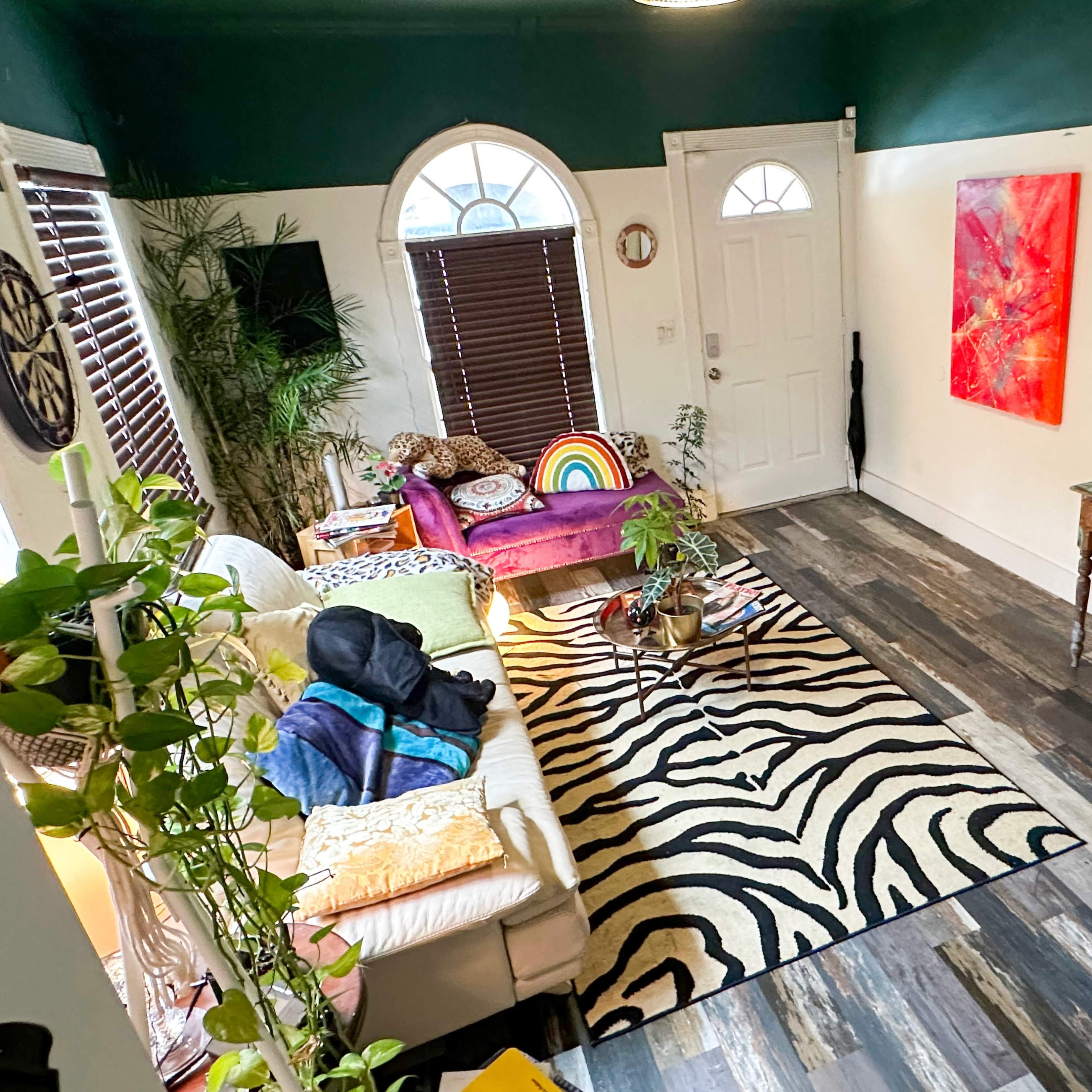 The room features a colorful seating area with a rainbow-striped cushion, a zebra-patterned rug, and a small desk with a monitor and chair against a vibrant green and white wall.