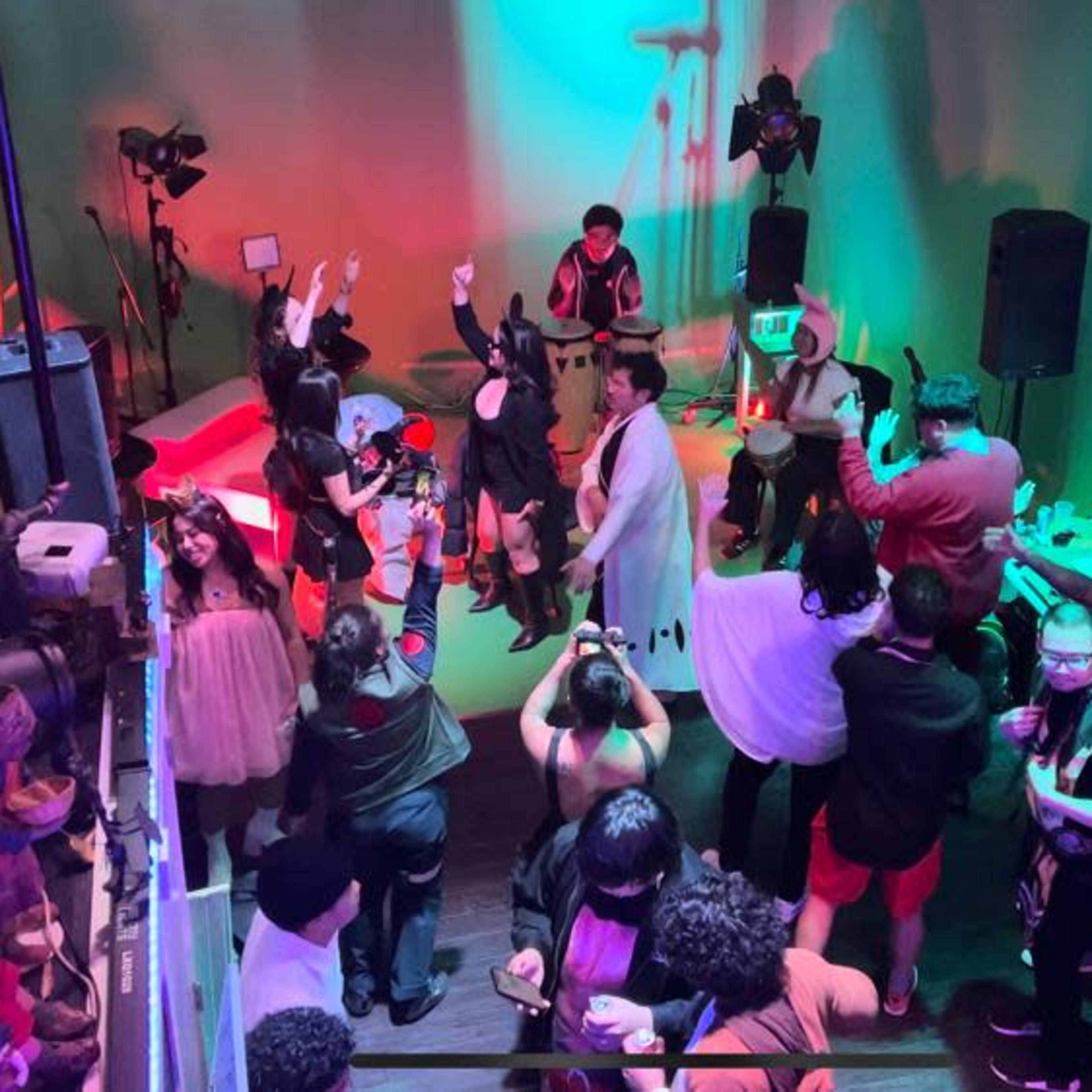 A large group of people gathers in a vibrant, dimly lit venue for a live performance, with cameras and lighting equipment positioned around the room.