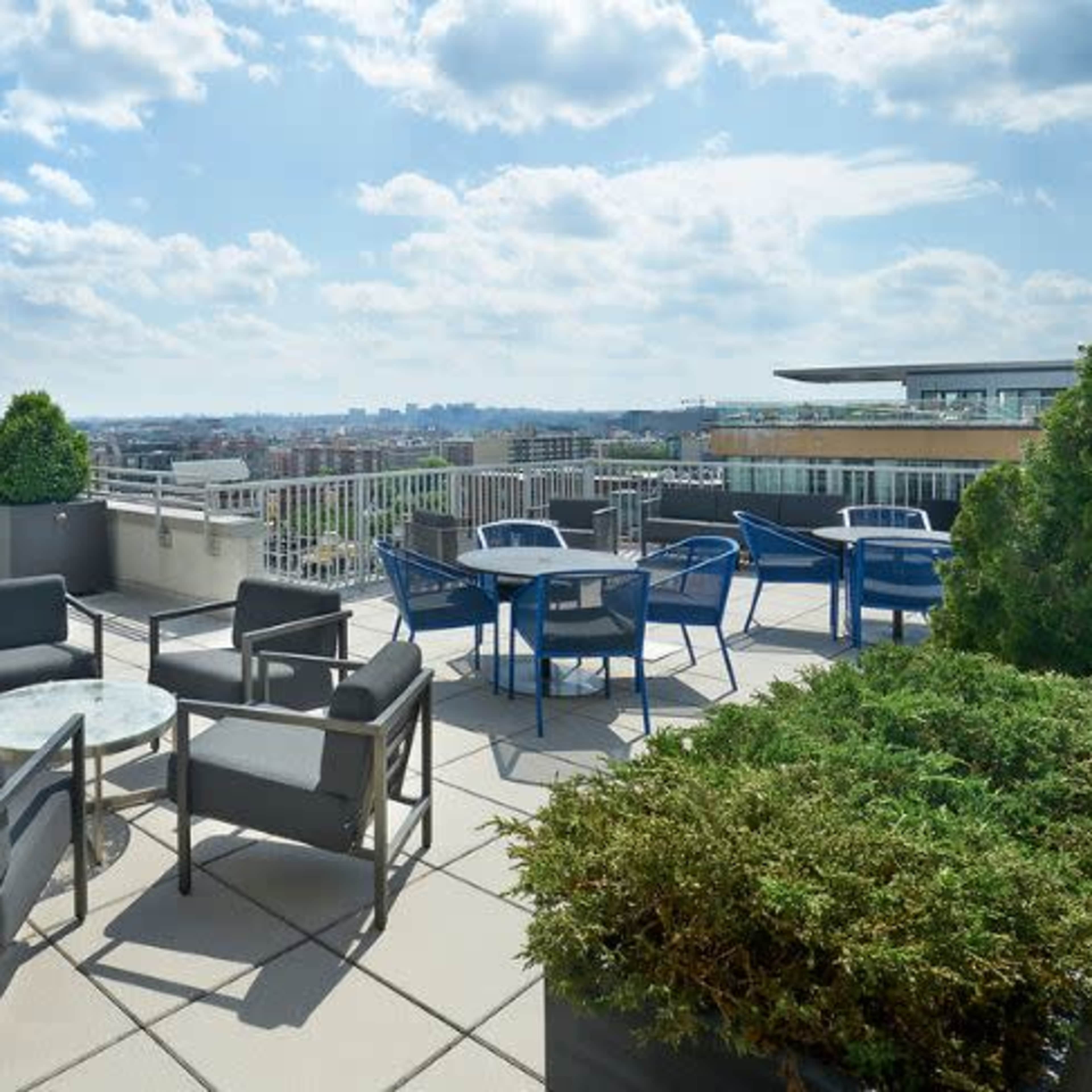 A rooftop terrace with seating arrangements, including chairs and tables, overlooking a city skyline under a partly cloudy sky.