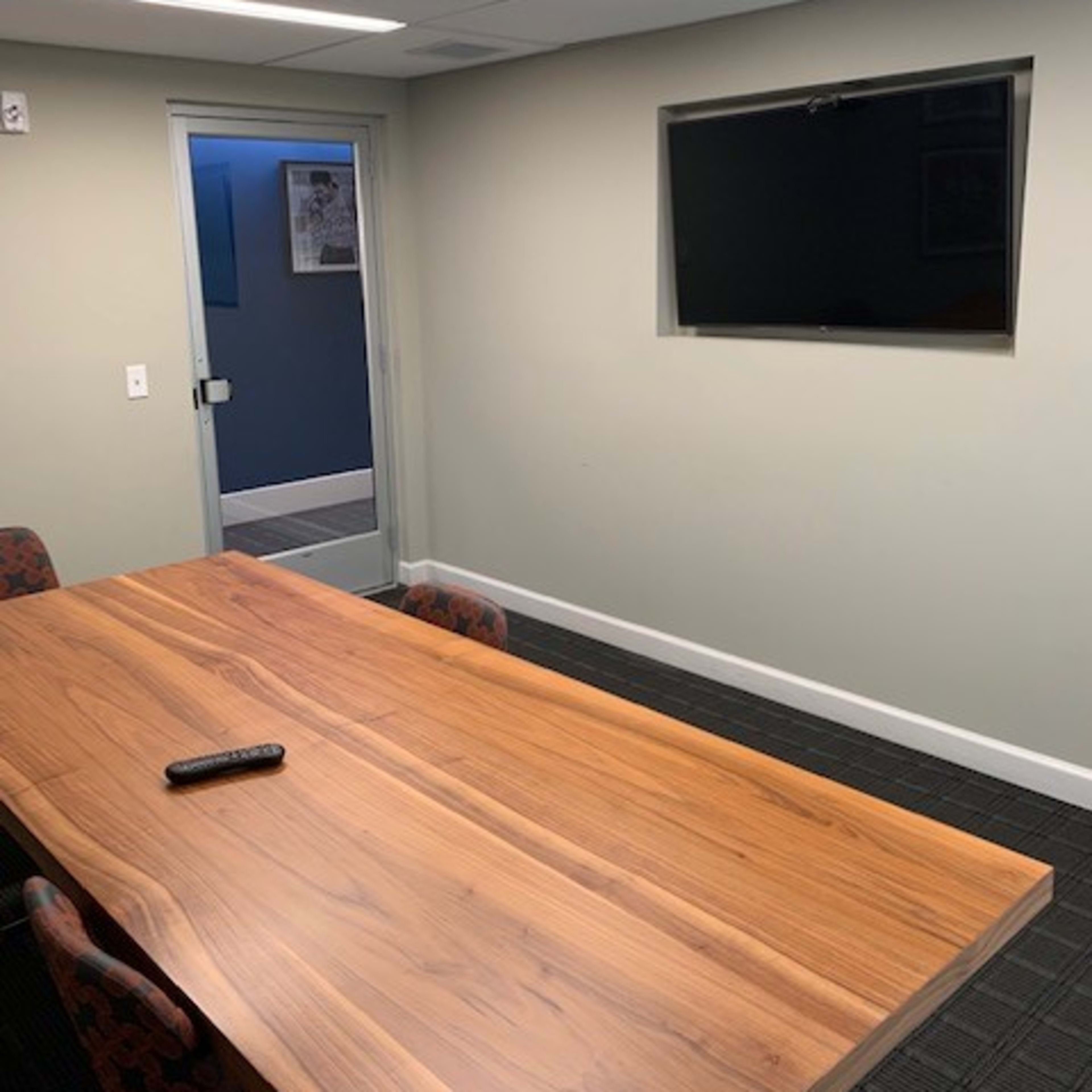 A conference room features a wooden table with a remote control on it, a mounted TV on the wall, and a glass door leading to another area.