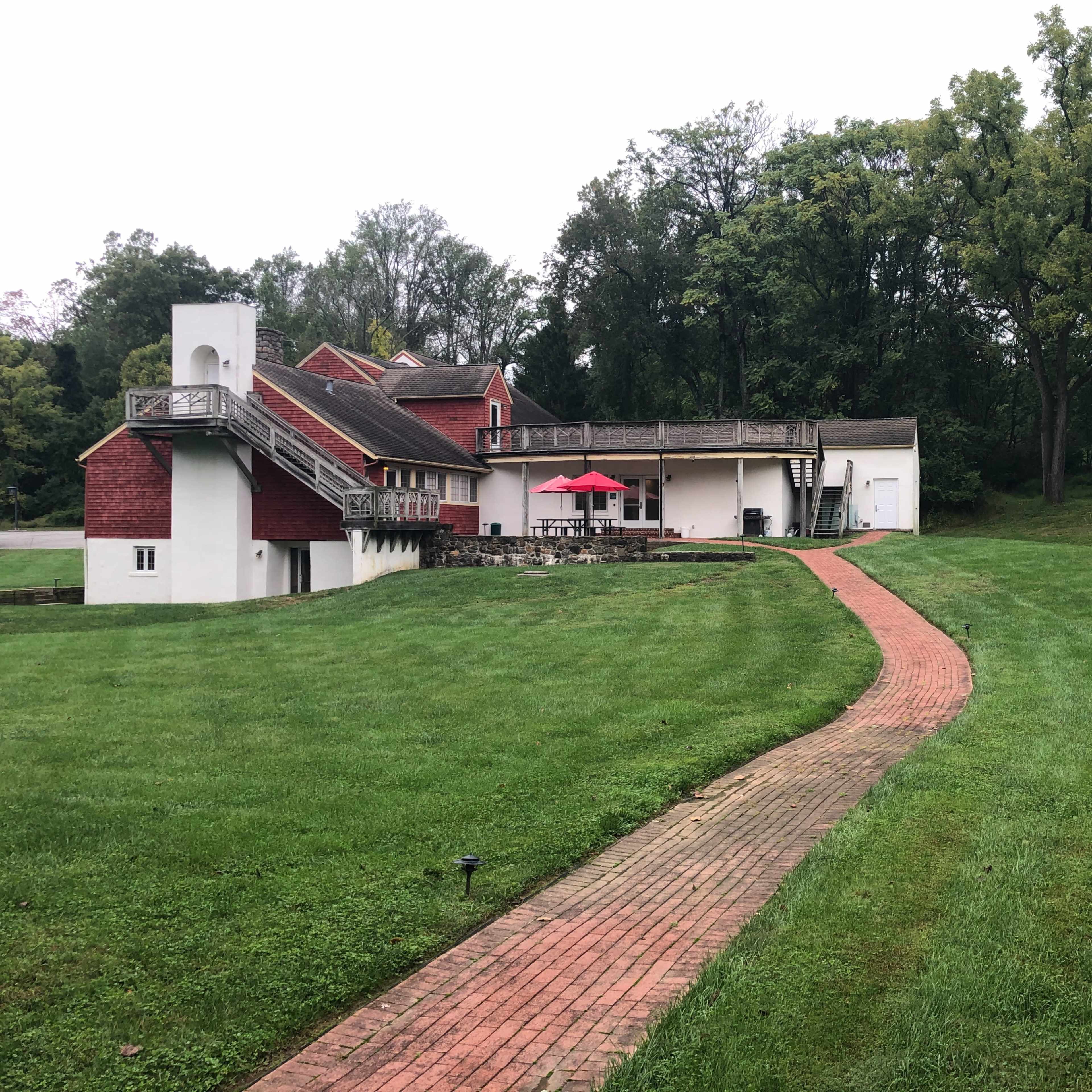 A brick pathway leads to a large, two-story building surrounded by greenery and trees, featuring an outdoor seating area with red umbrellas.
