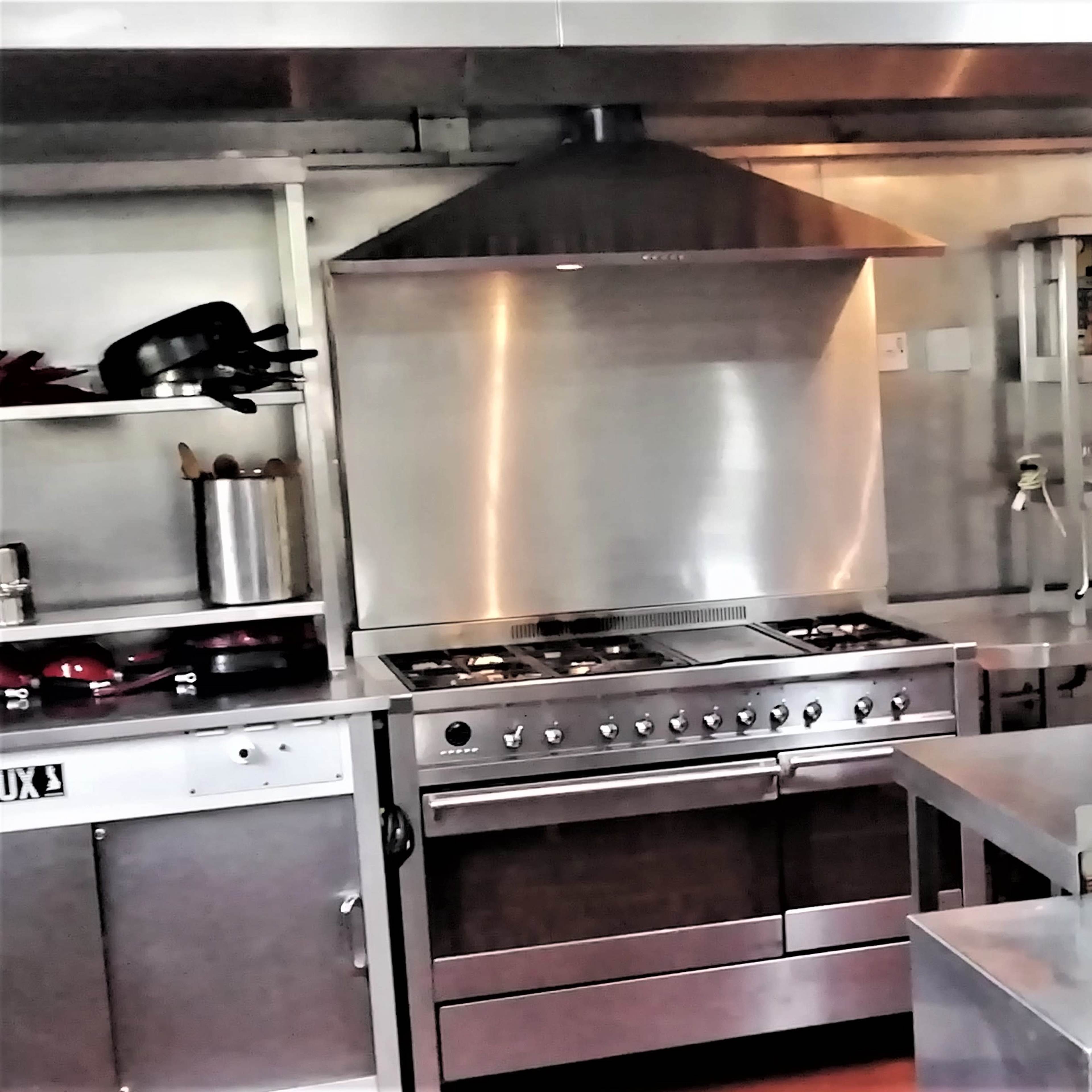 A stainless steel kitchen with a gas stove, an exhaust hood, and various kitchen utensils and appliances on the shelves.