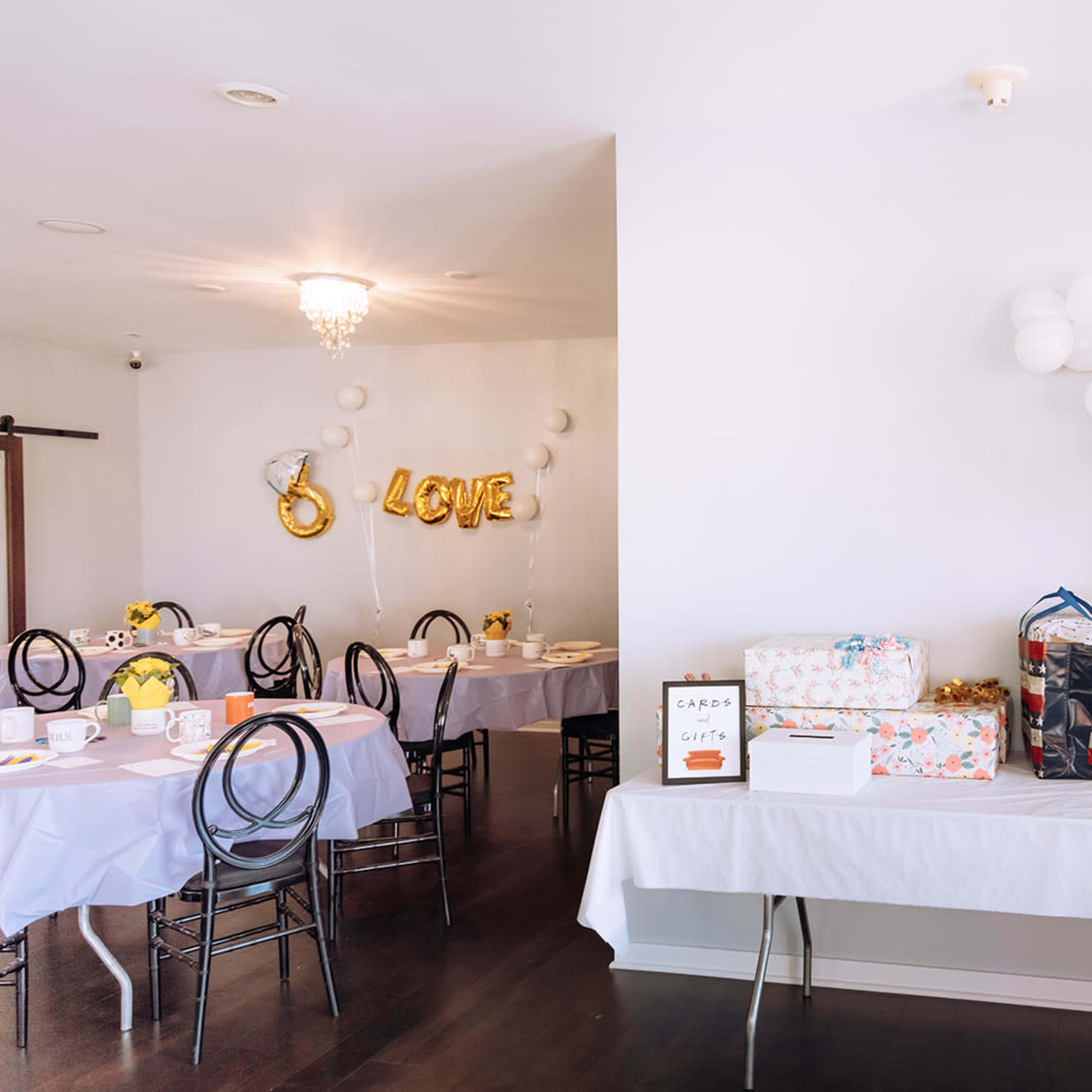 A dining area features tables set with white tablecloths and a gift table adorned with colorful wrapped presents and balloons.