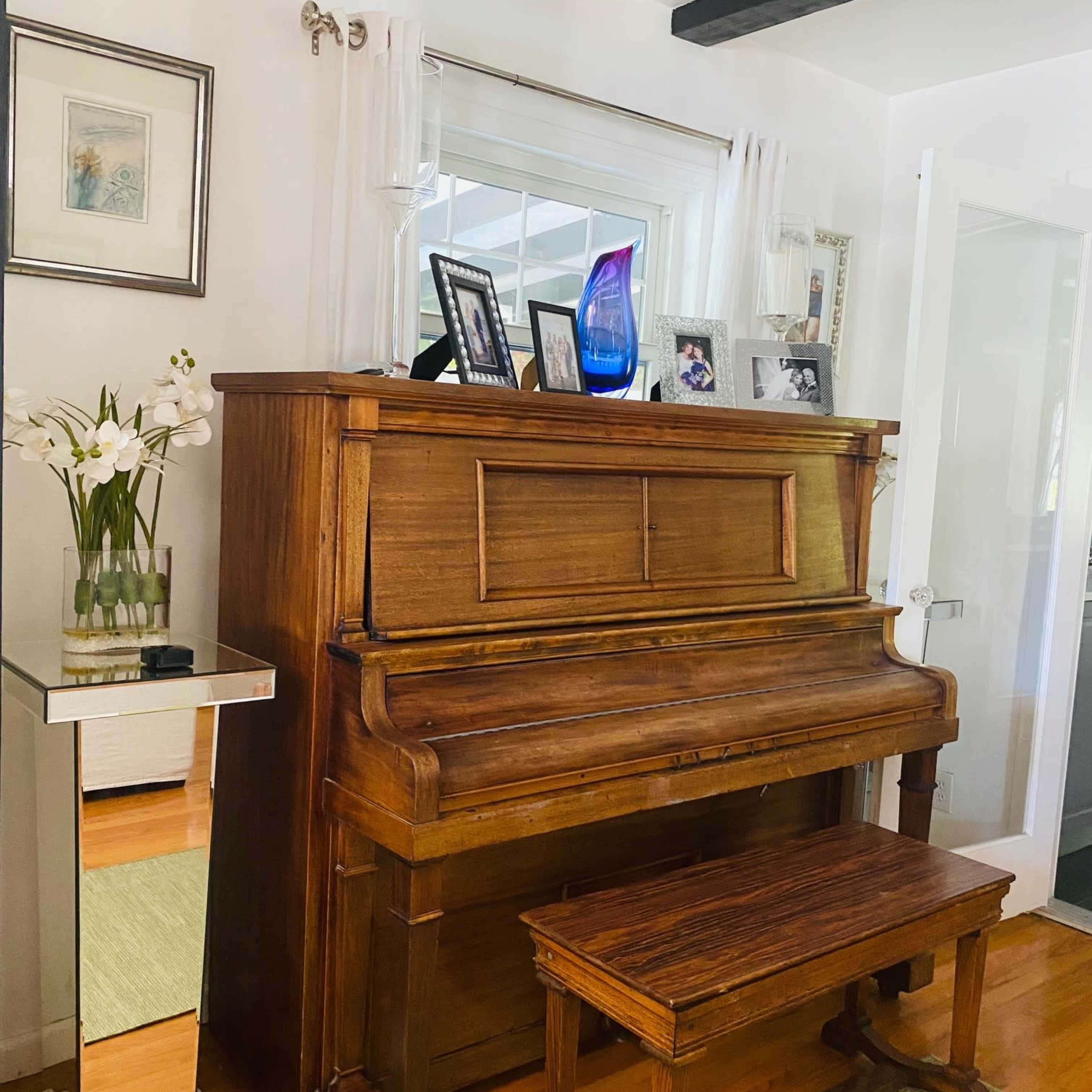A wooden piano is placed against a wall, accompanied by a matching bench, a mirrored side table, and decorative photographs on display.
