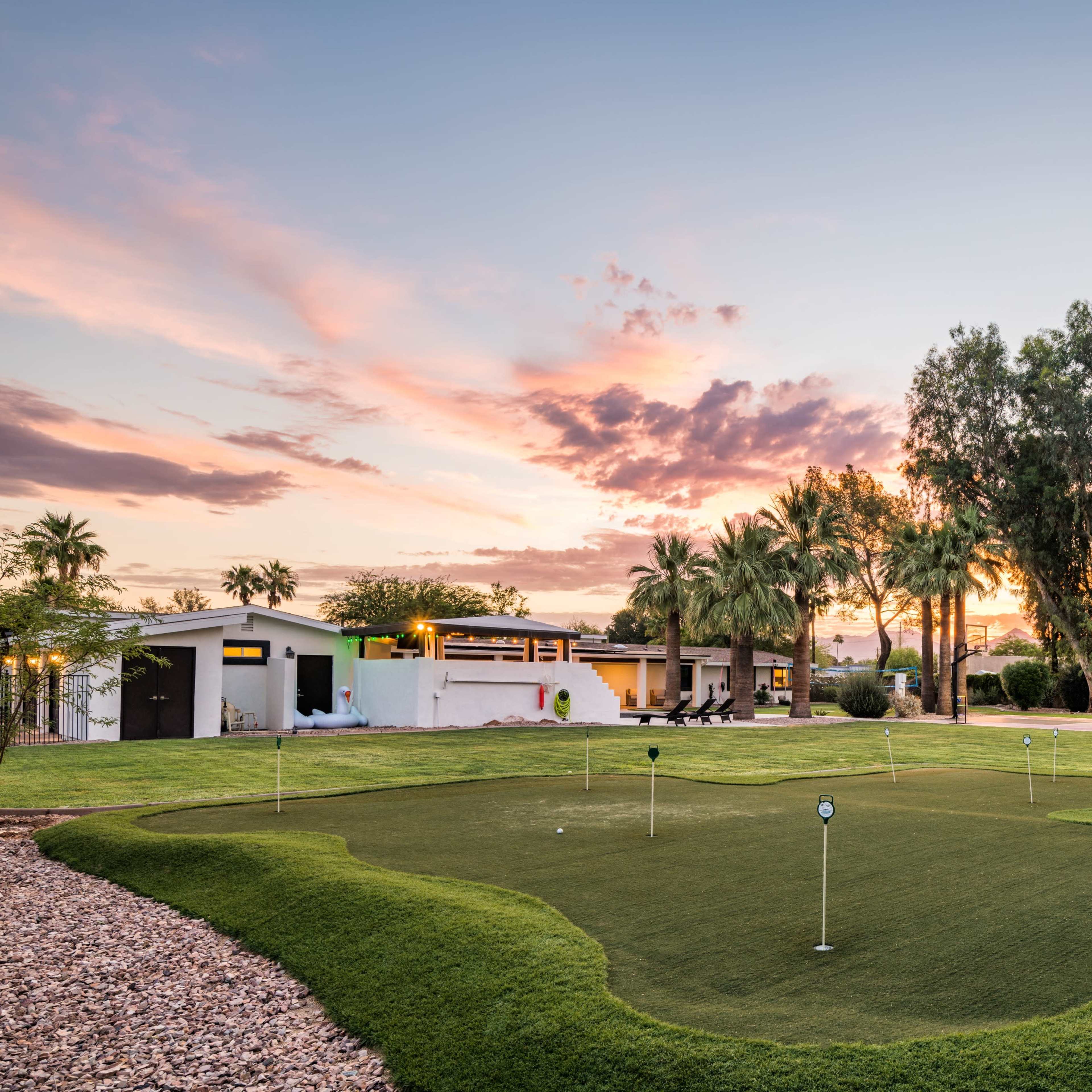A well-maintained putting green is surrounded by grass and palm trees under a sunset sky.