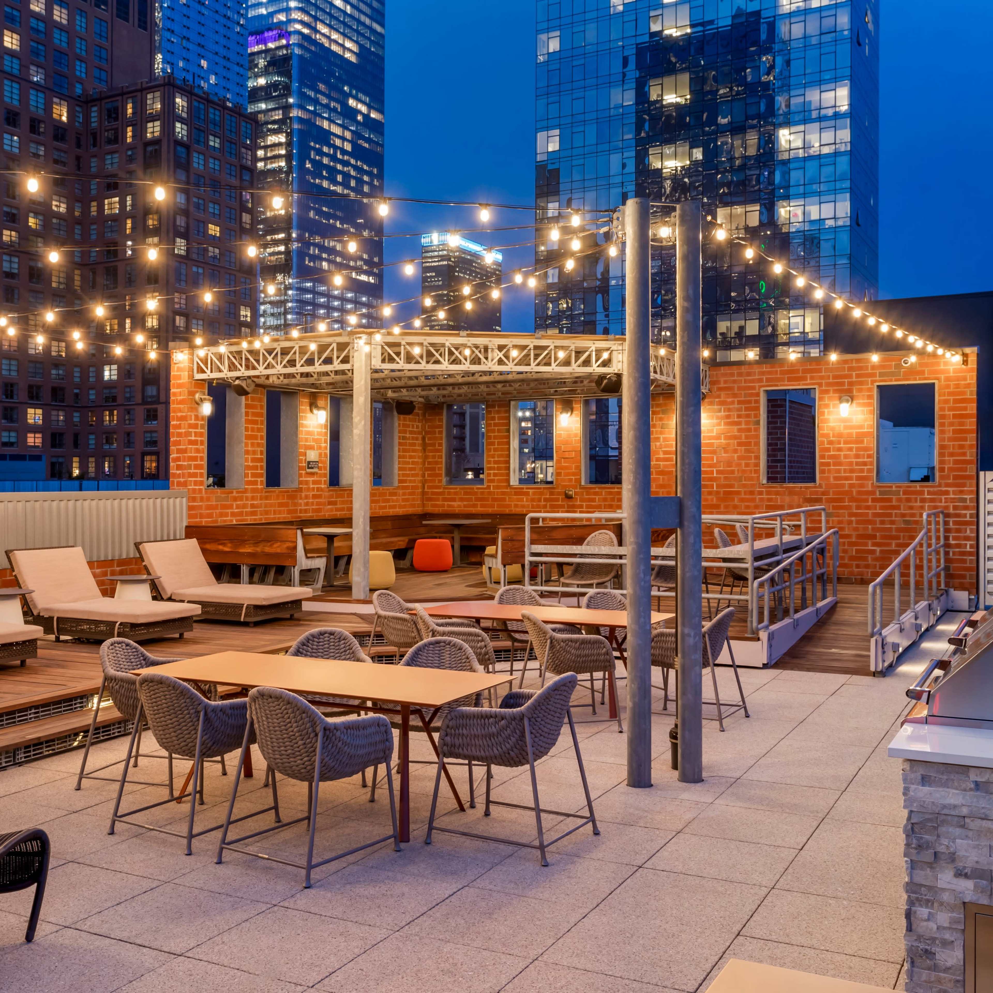 The rooftop terrace features a seating area with lounge chairs and tables, illuminated by string lights against a backdrop of skyscrapers at dusk.