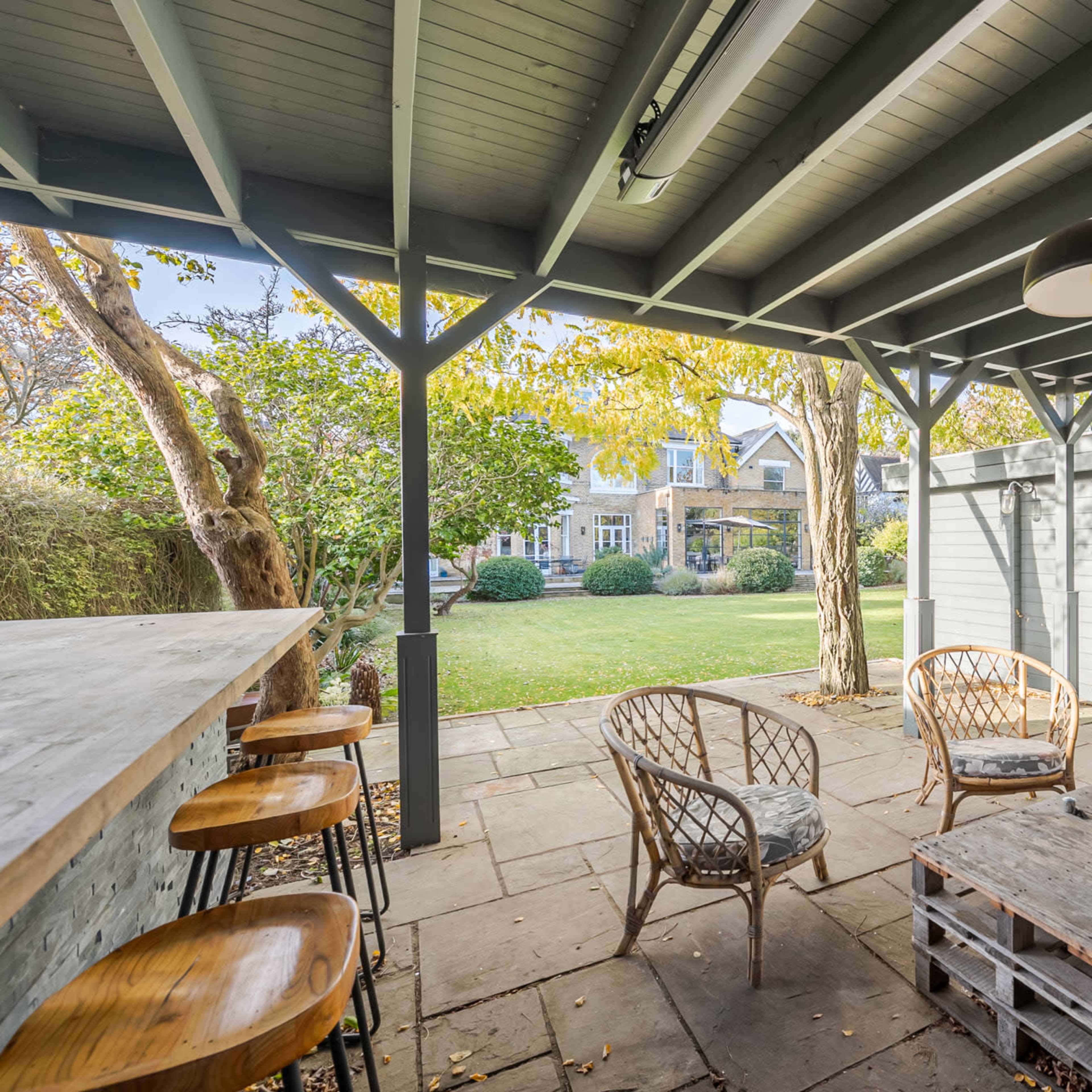 A covered outdoor seating area with a wooden bar, rattan chairs, and a table, overlooking a grassy backyard with trees.