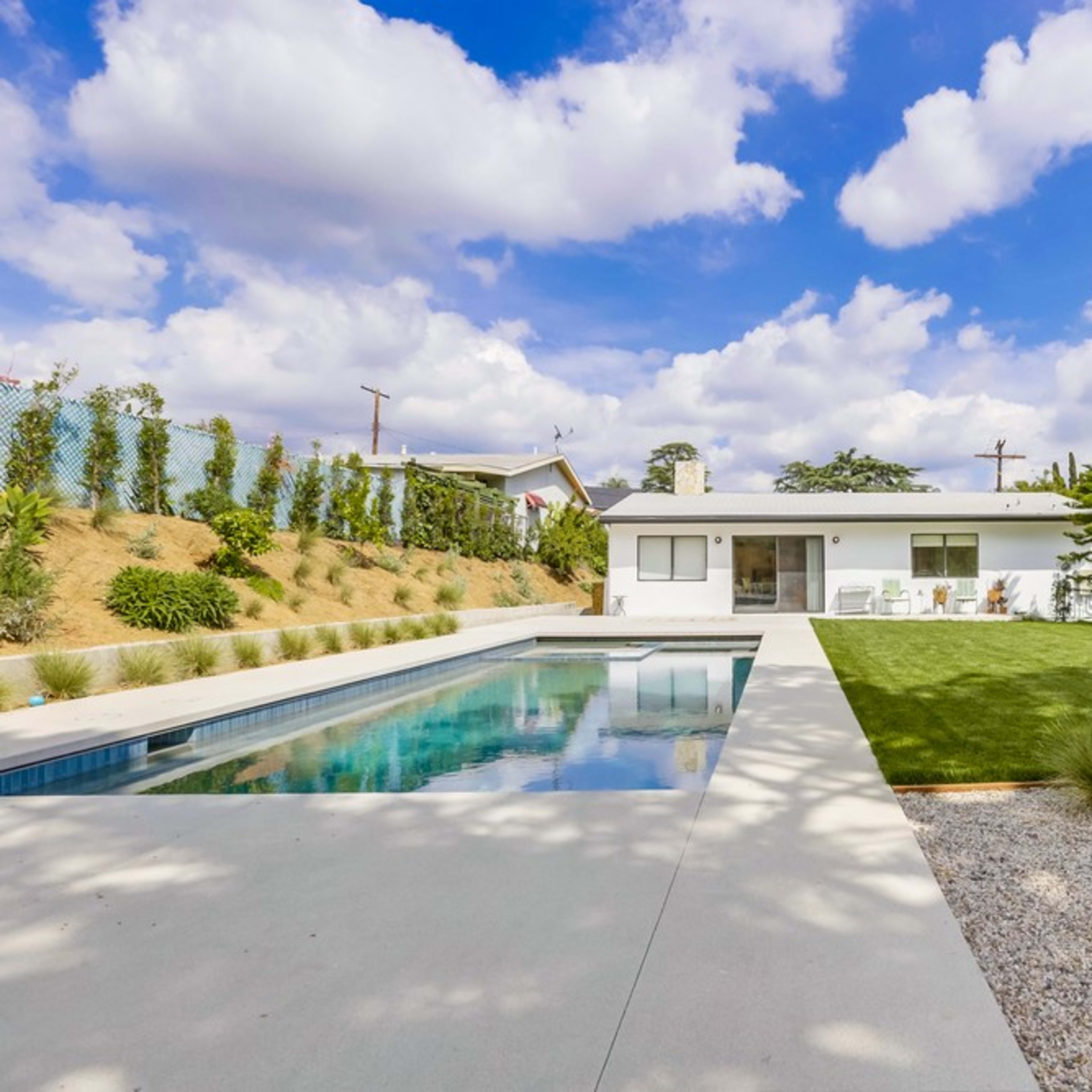 A modern house with a pool in the foreground, set in a landscaped yard that features green grass and decorative plants under a partly cloudy sky.