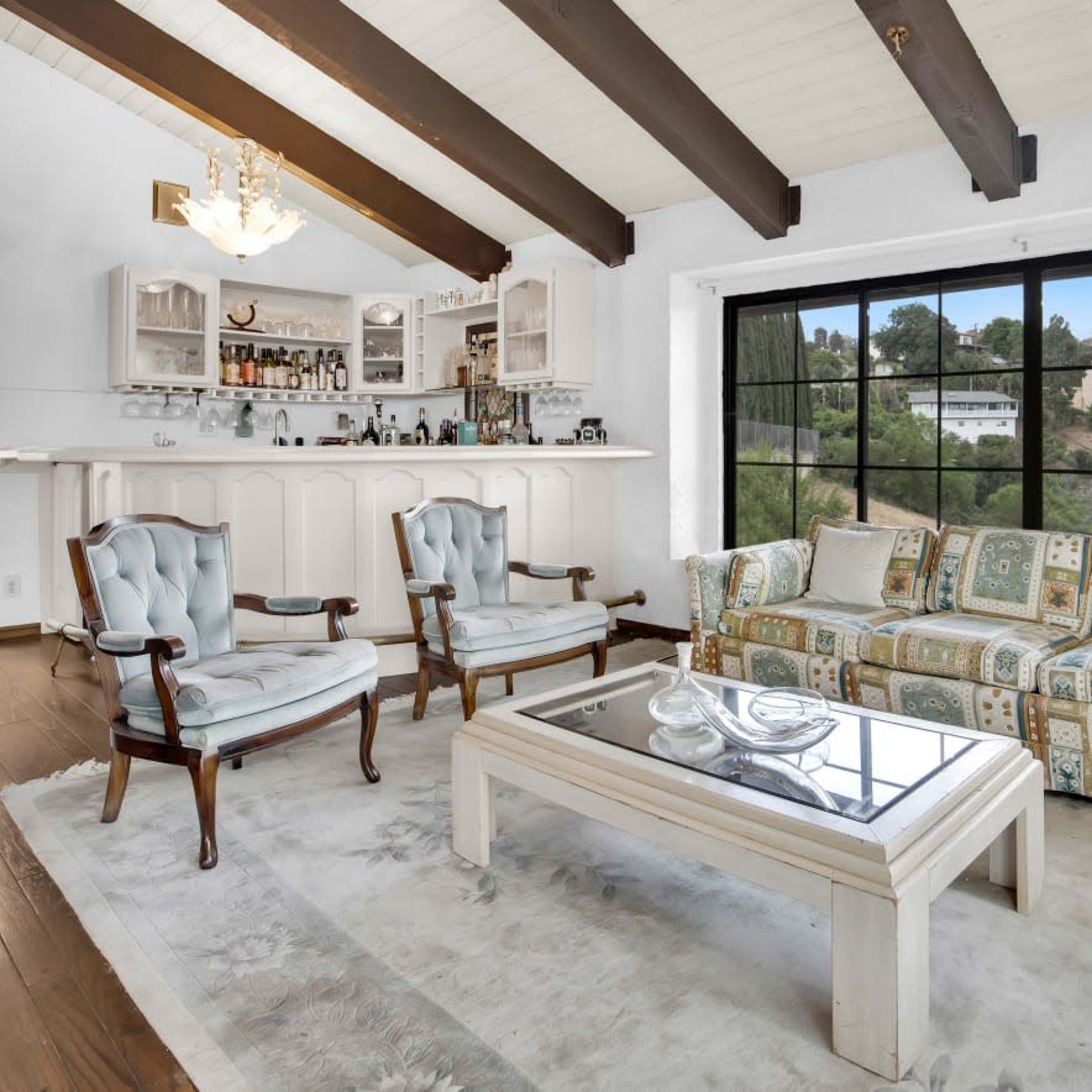 The living room features a bar area, a patterned sofa set, and large windows overlooking a green hillside.
