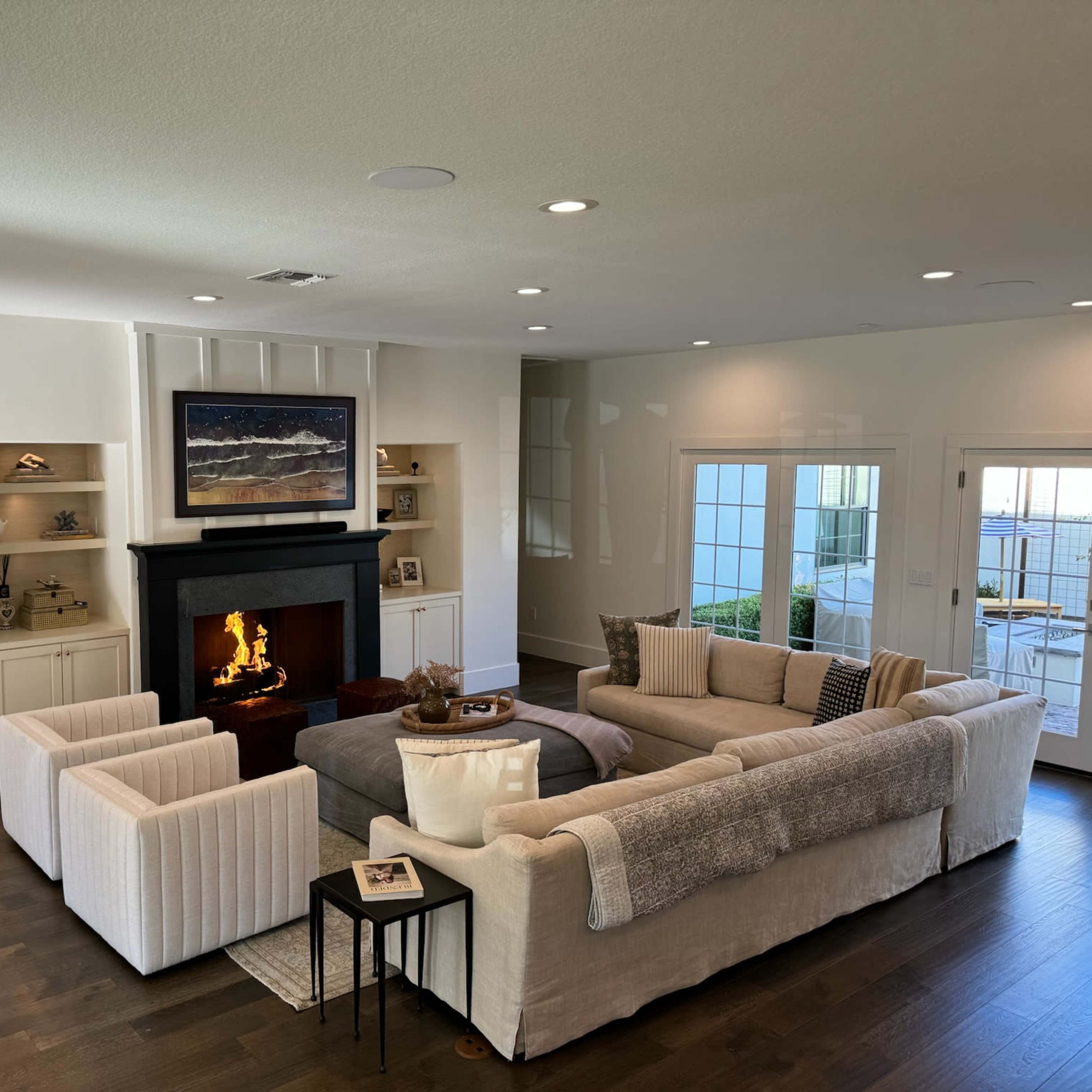 A modern living room with a large sectional sofa, two armchairs, a fireplace, built-in shelves, and large windows offering views of the outdoors.