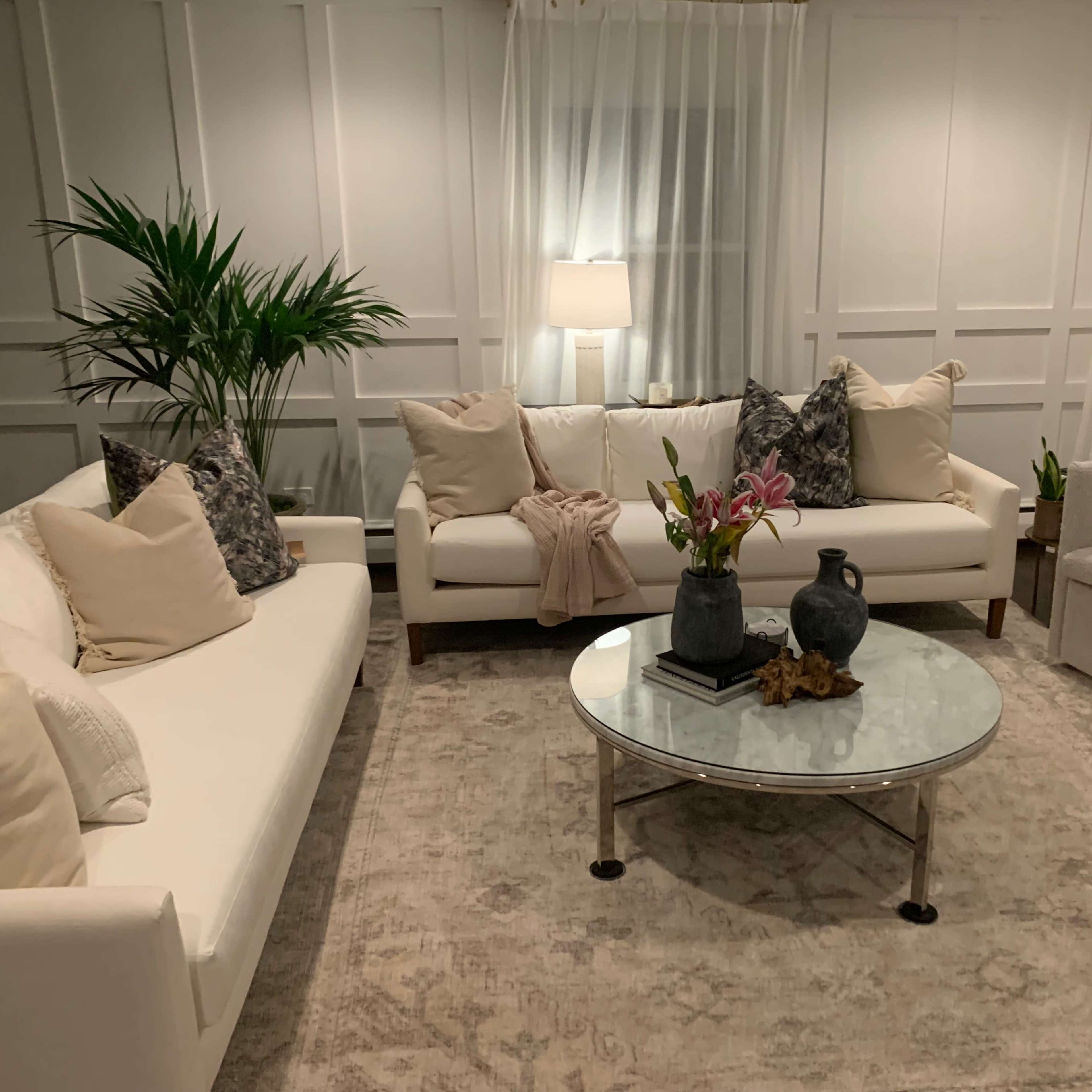 A modern living room features white sofas, a glass coffee table, and decorative plants under soft lighting.