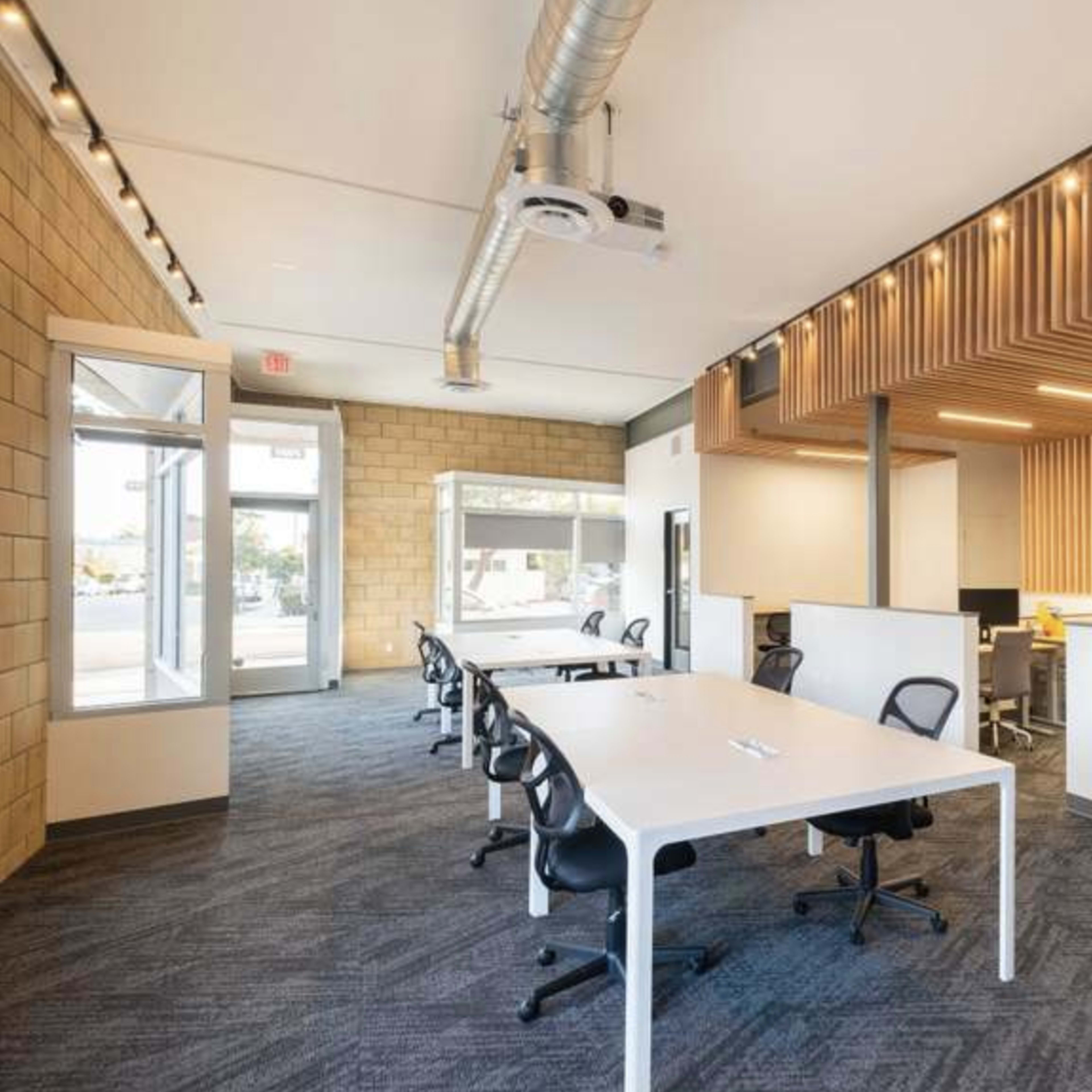 A modern office space featuring several white desks with black chairs, large windows, and a mix of wood and concrete wall textures.