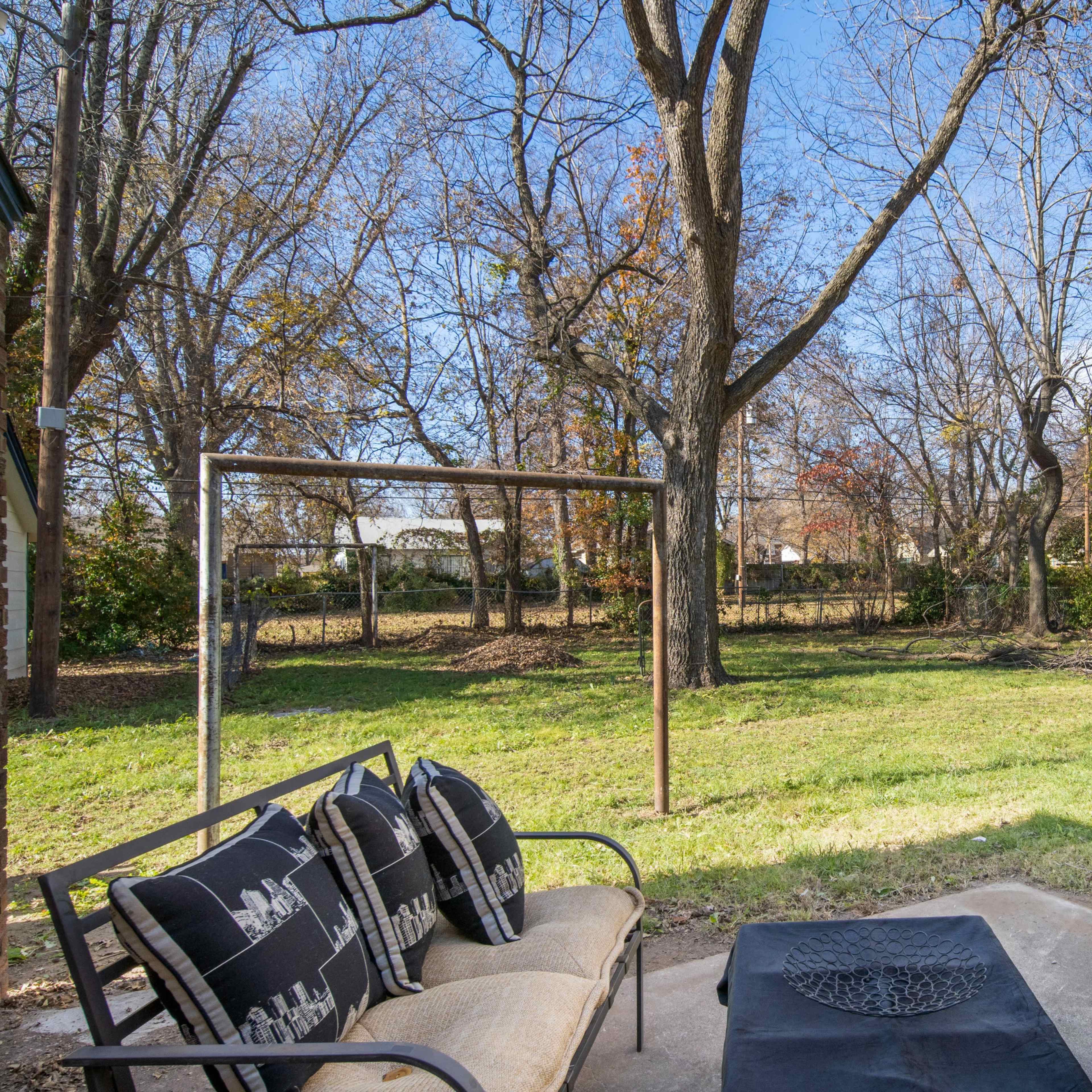 A backyard scene features a seating area with black upholstered furniture and a small table, surrounded by trees and a grassy lawn.